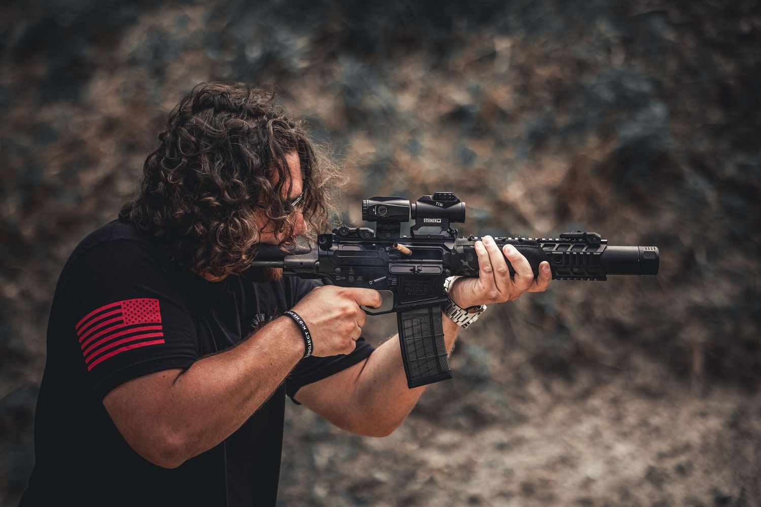 Person in black t-shirt with red flag patch aims AR-style rifle with scope and silencer outdoors.