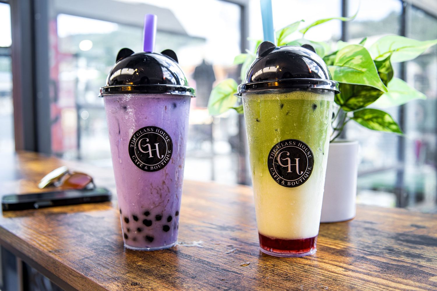 Two bubble tea drinks in clear plastic cups on a wooden table, one purple taro and one yellow with red syrup at bottom.