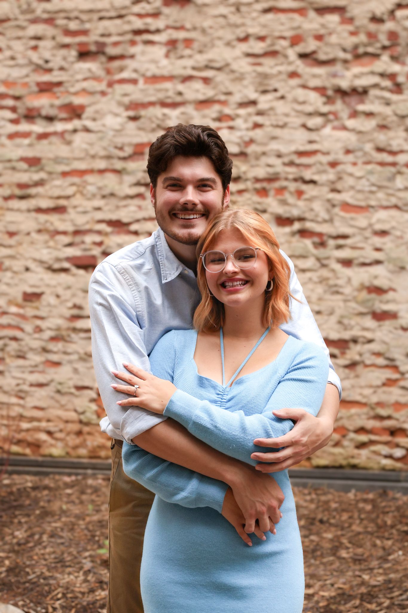 Happy couple embraces in front of weathered brick wall during engagement photoshoot.