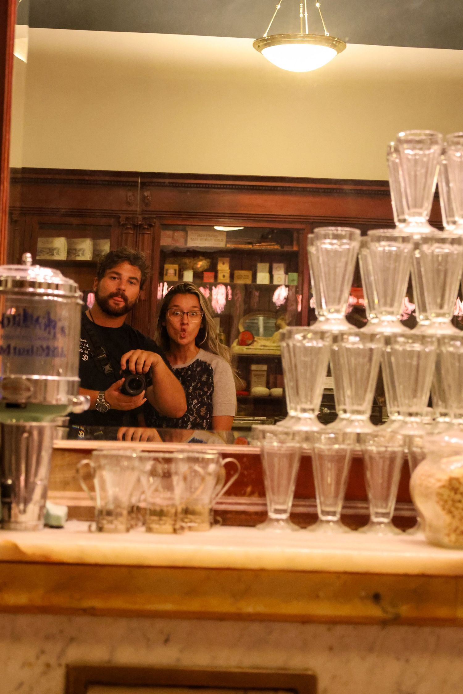 Behind a bar counter with stacked glasses, people with a camera stand next to a professionally arranged display.