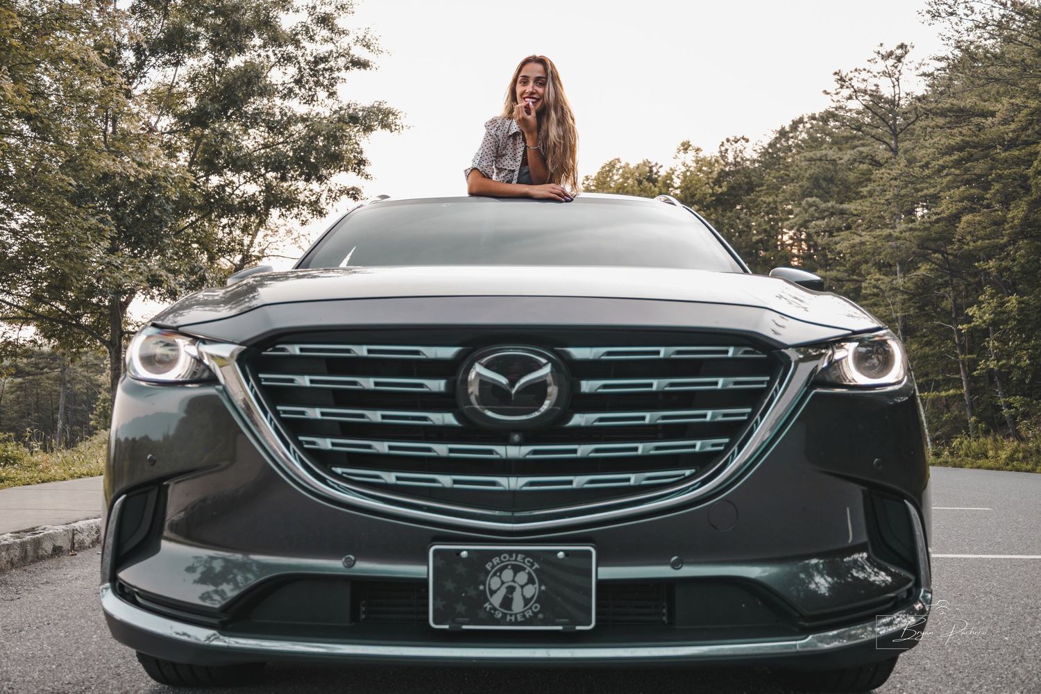 Sleek gray Mazda CX-9 SUV parked on road with forest background and someone sitting on hood.