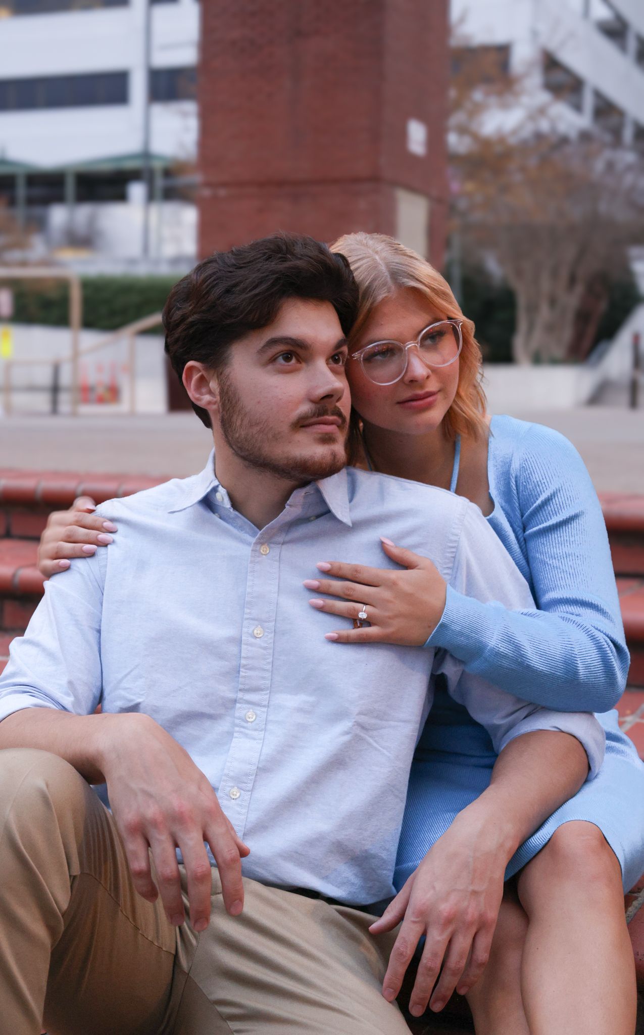 A couple wearing casual outfits embrace while sitting outdoors, with a brick building visible in the background.