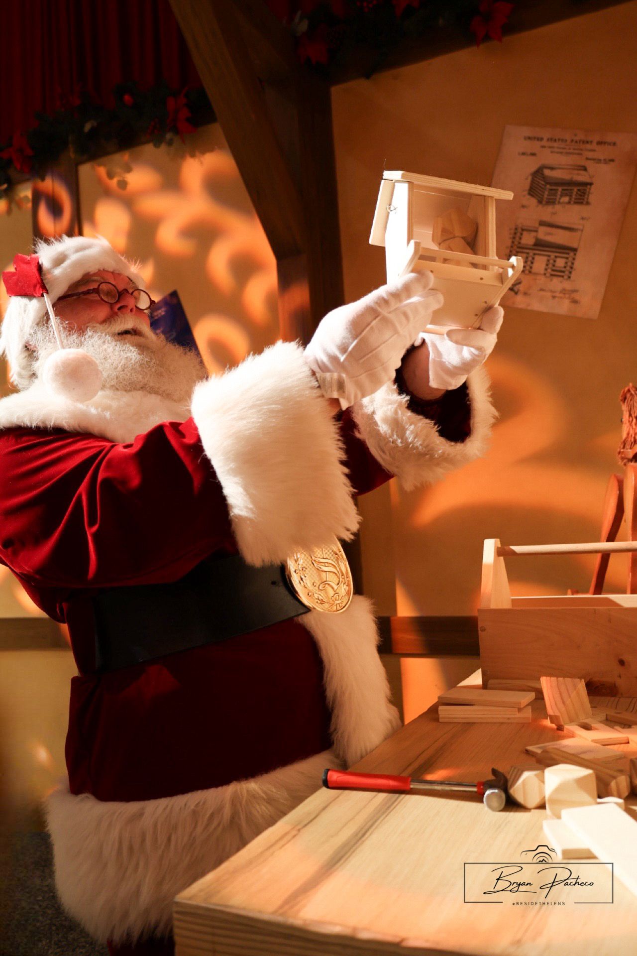 Santa Claus wearing traditional red suit and white gloves examines paperwork at a desk with an orange-lit background.