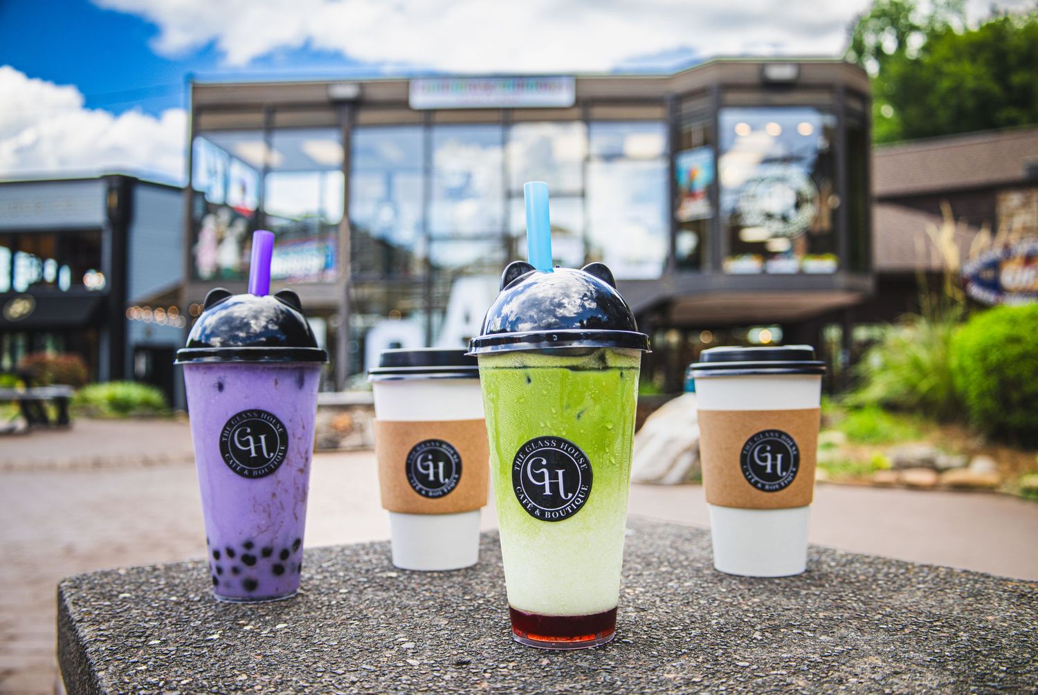 Four colorful bubble tea drinks from The Glass House cafe arranged on a concrete ledge with modern buildings in background.