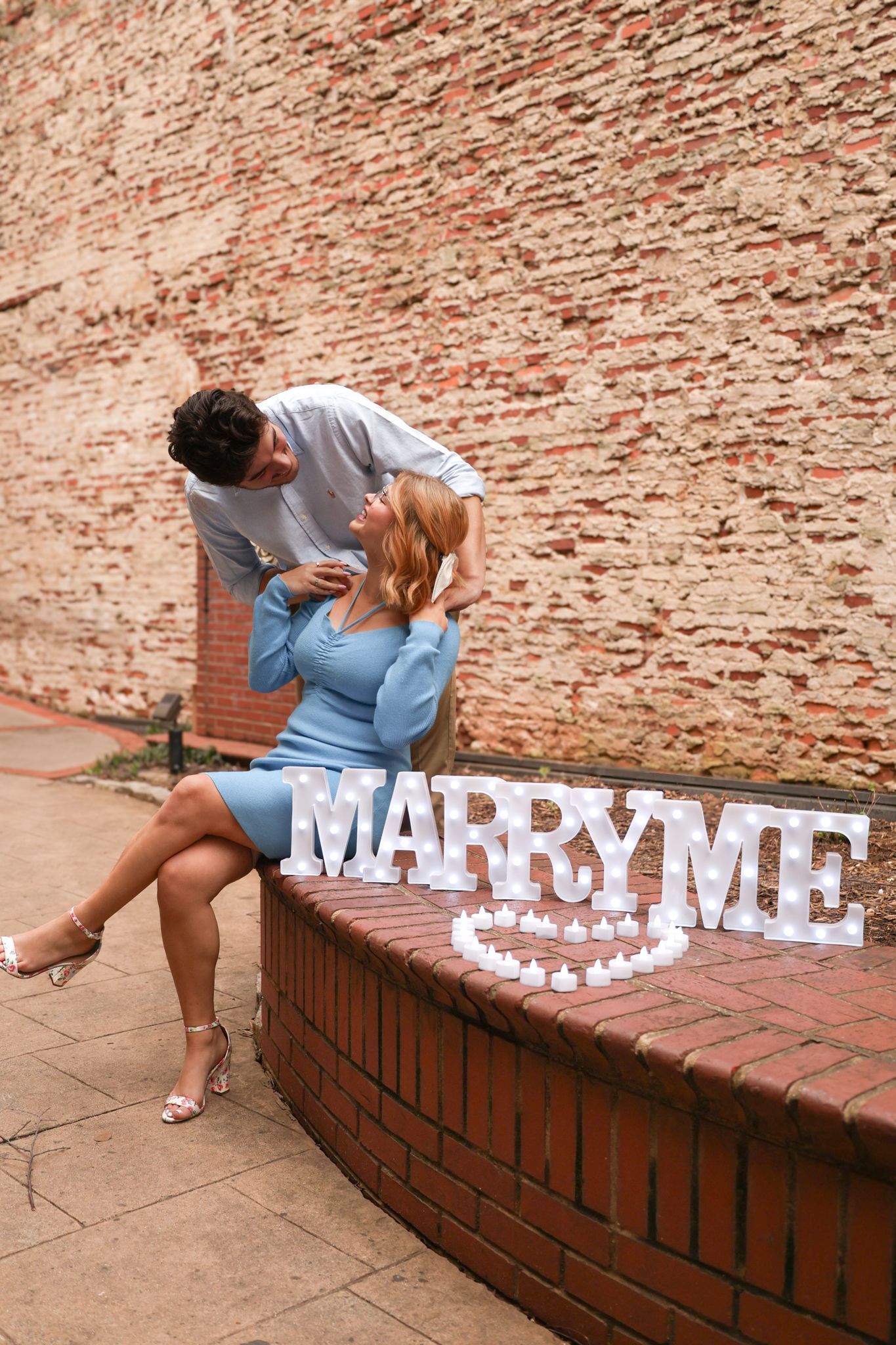Marriage proposal scene with 'Marry Me' text display on brick wall ledge.