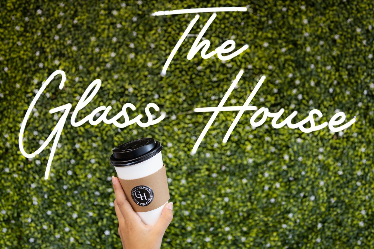 The Glass House logo displayed in white cursive text on a green hedge wall with a branded drink cup.