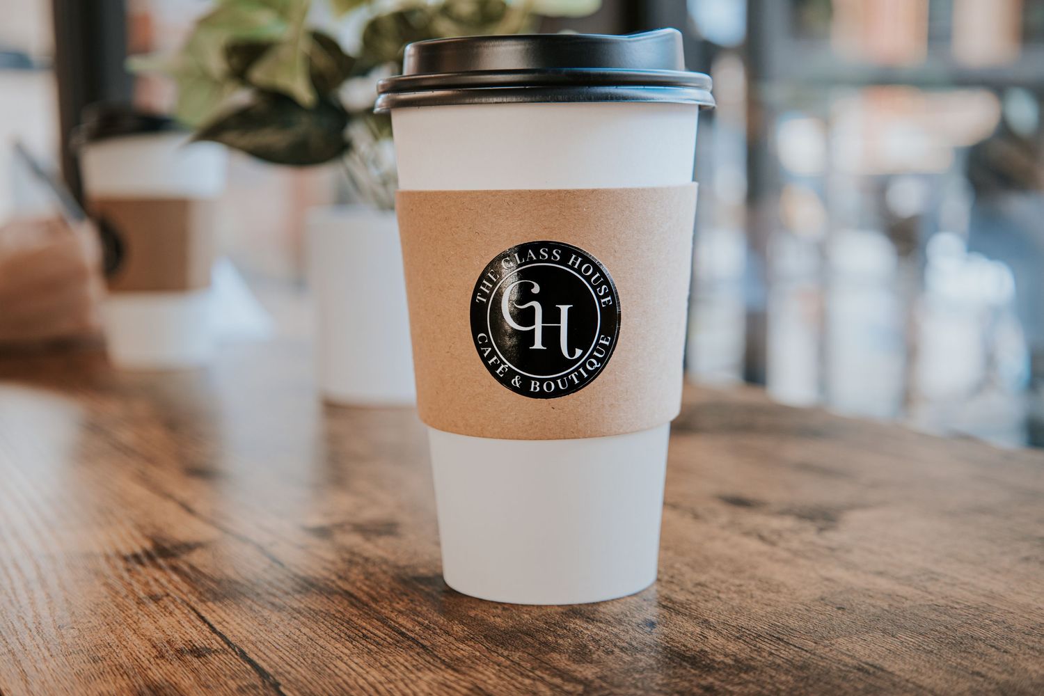 A white coffee cup with GH logo branding sits on a wooden table in a cafe setting.