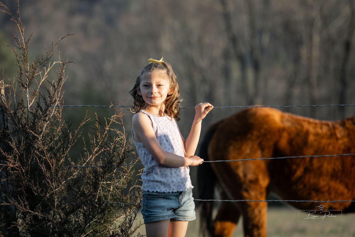 Young person in denim shorts and white top stands in a field with a brown horse at sunset.
