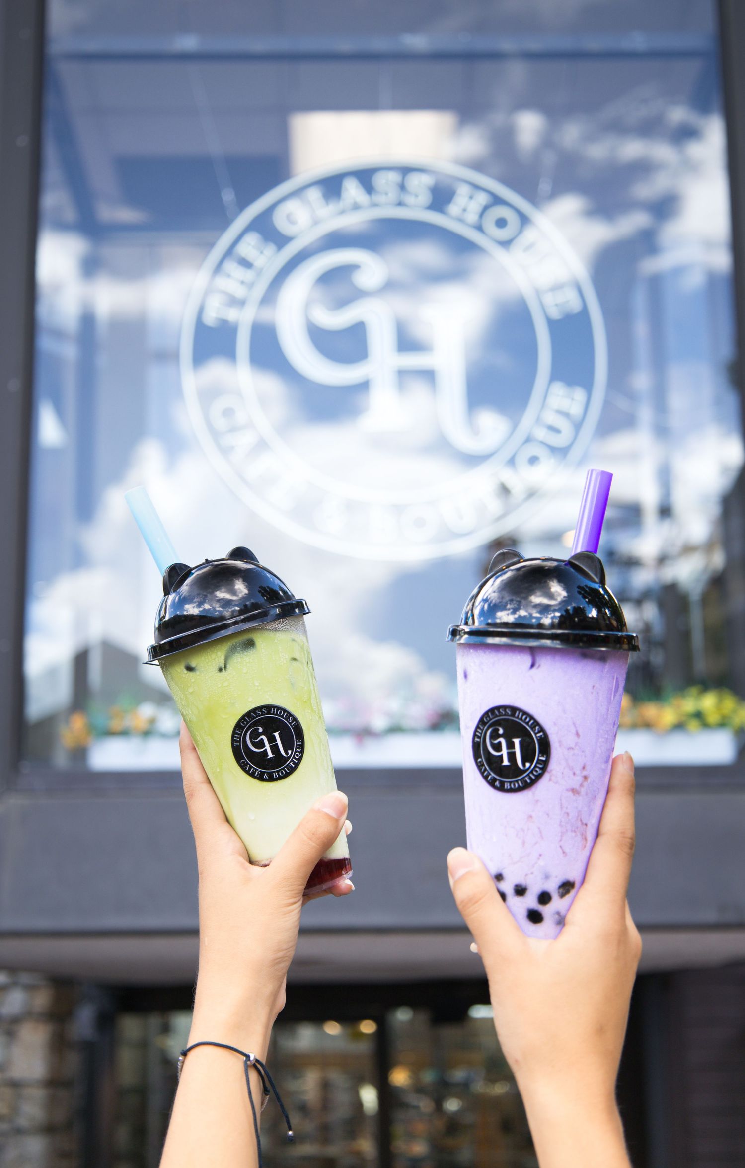 Two hands holding colorful bubble tea drinks with the Glass House logo visible in the window background.