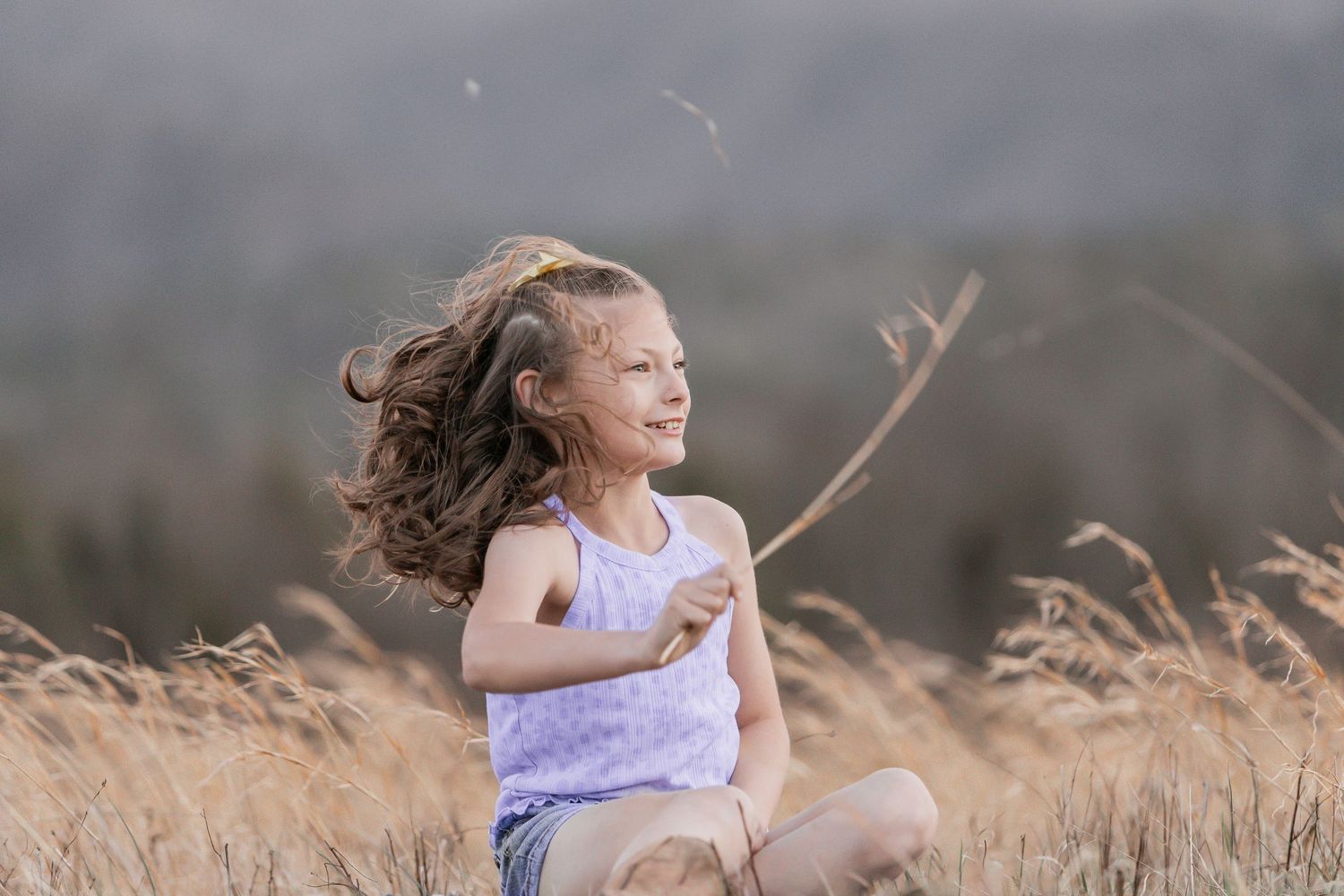 A young person sits joyfully in a golden wheat field with windswept hair on a sunny afternoon.
