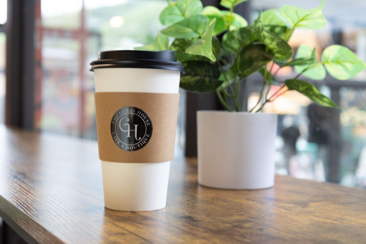 A coffee cup with a cafe logo sits on a wooden table next to a white planter with leafy green plants.