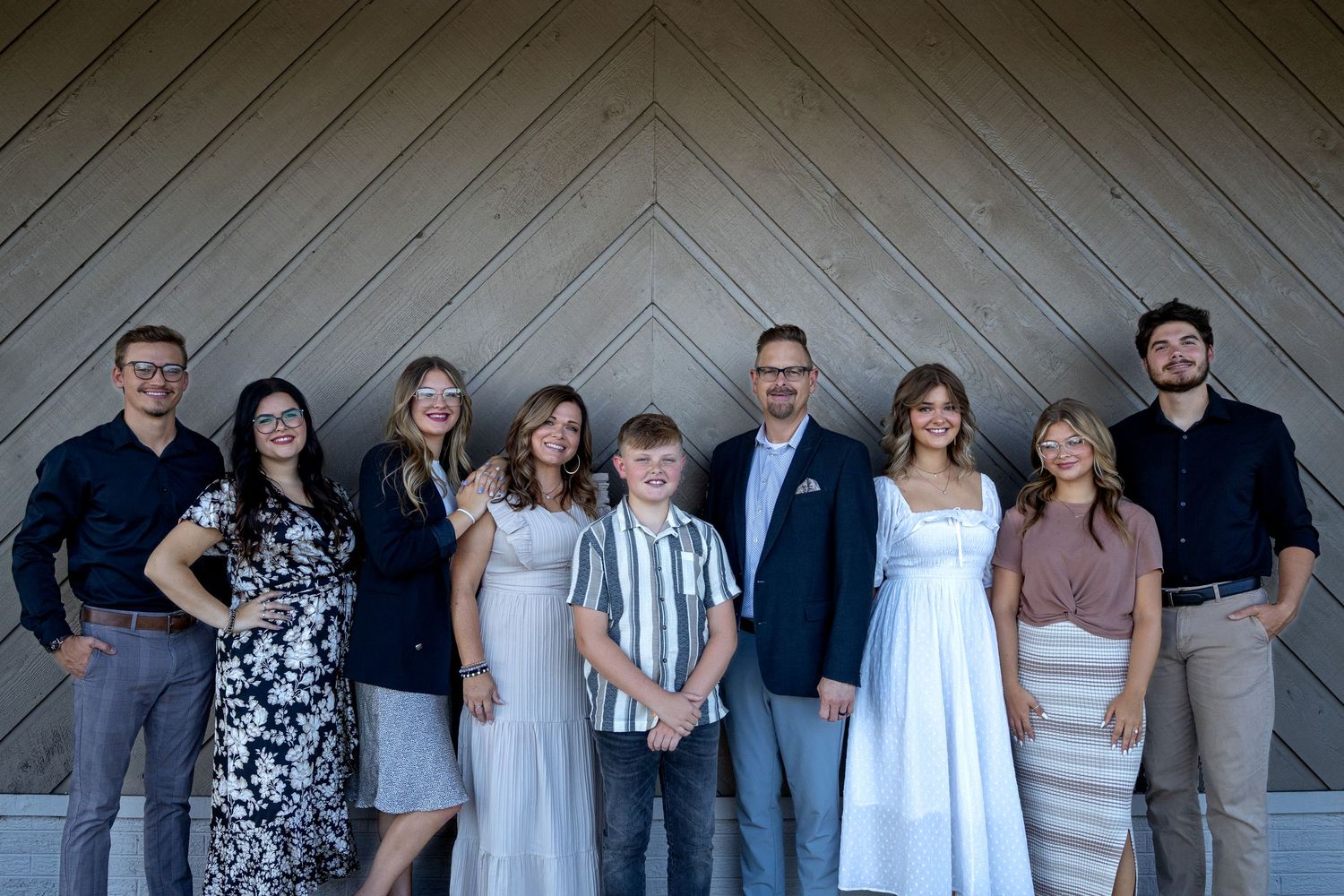 A group of well-dressed people stand together against a wooden wall with chevron pattern siding.