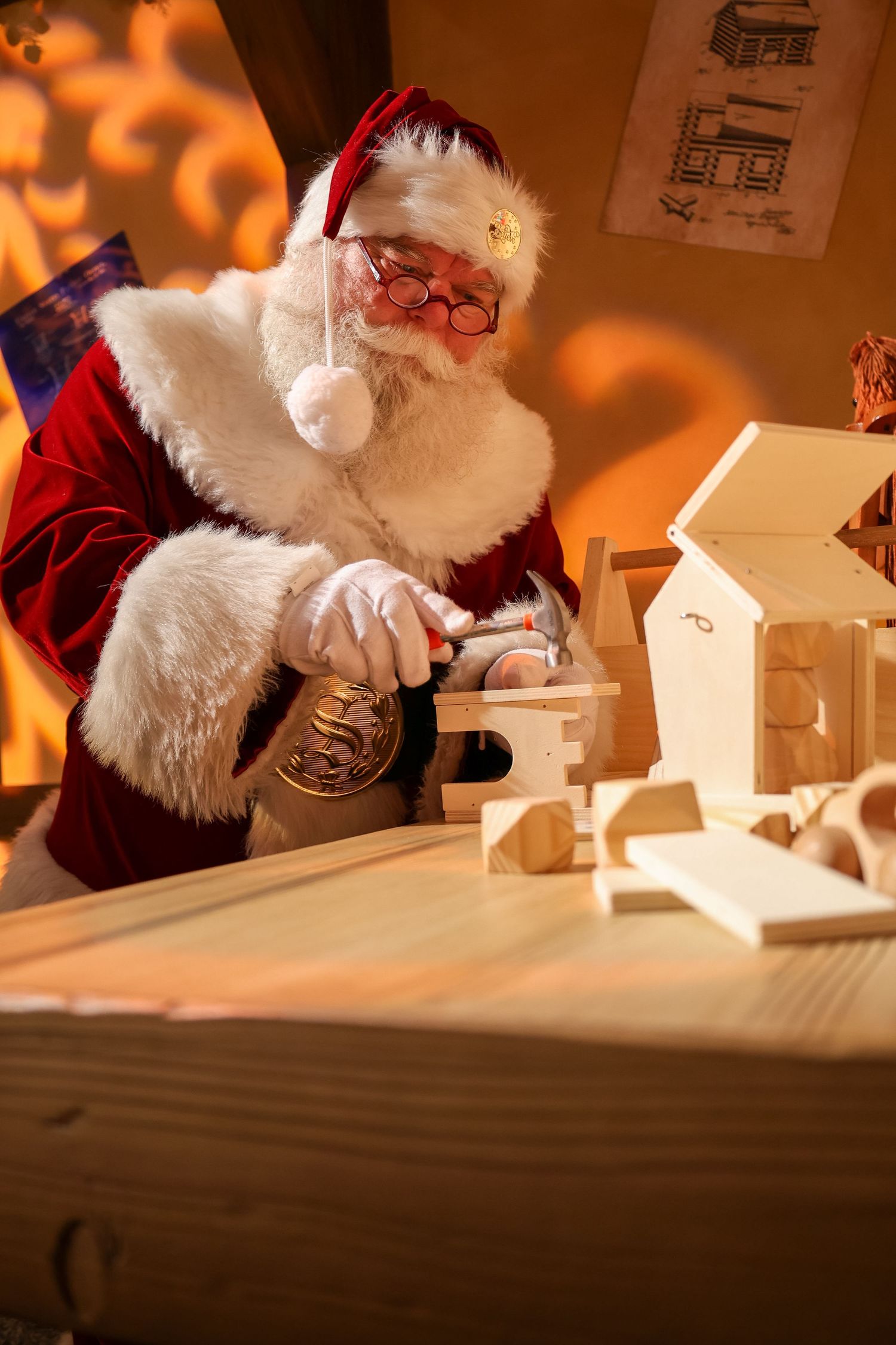 A festive workshop scene showing someone in a red and white suit building a wooden toy house in warm lighting.