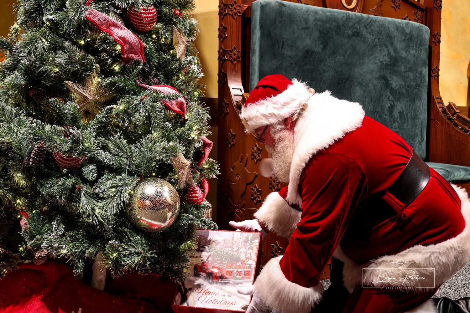 A decorated Christmas tree beside a traditional holiday figure in red suit writing on paper while seated.