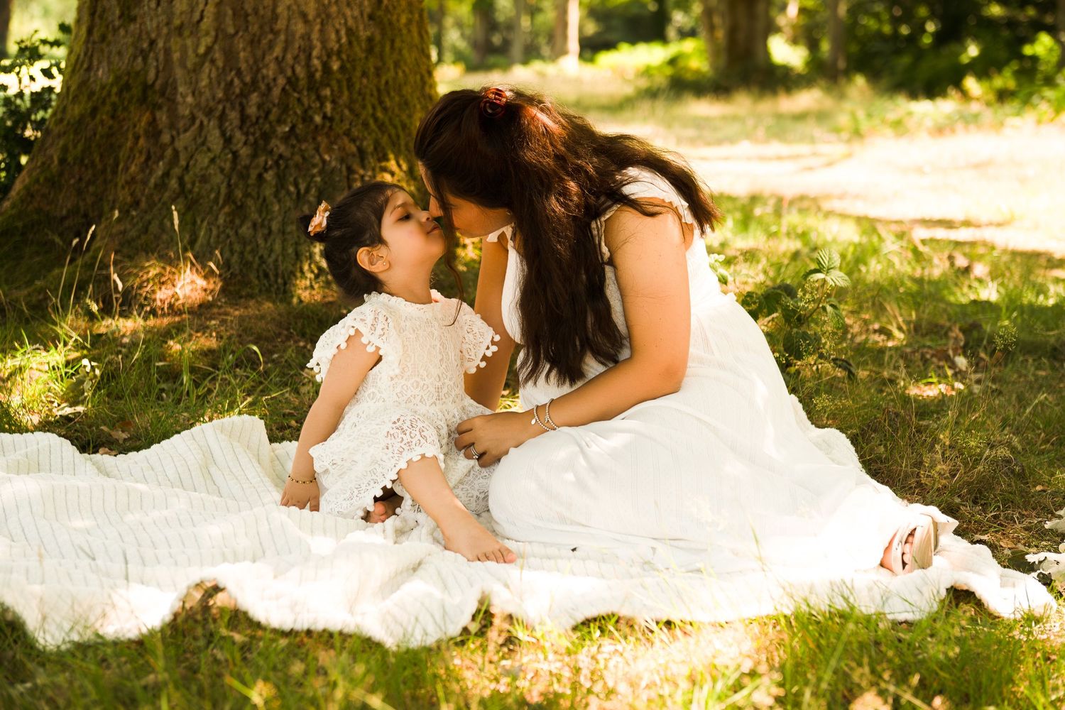 Family wearing white outfits sits on blanket in dappled sunlight during outdoor photo session.