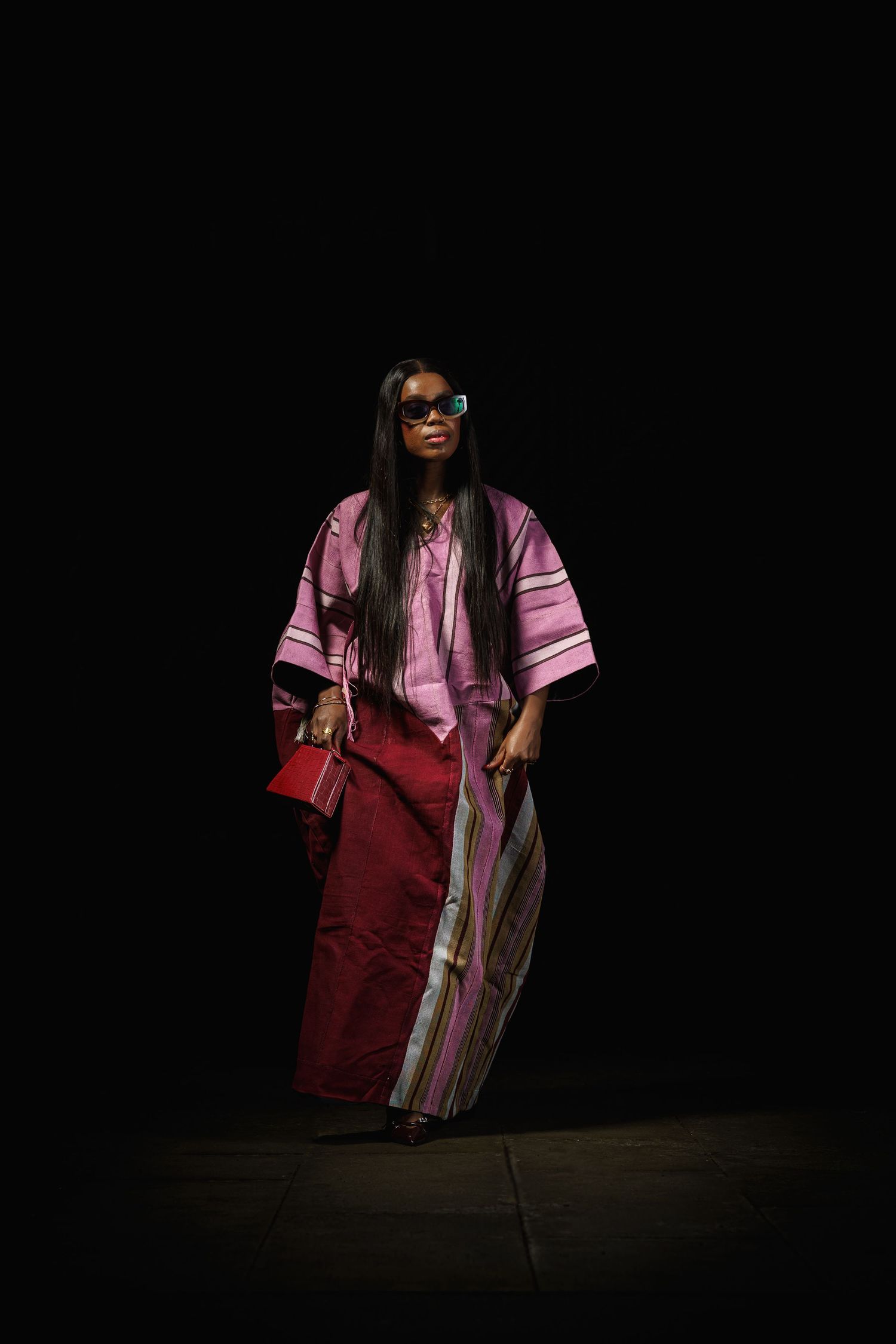 Model walks runway in pink and burgundy striped maxi dress with sunglasses and red handbag against dark background.