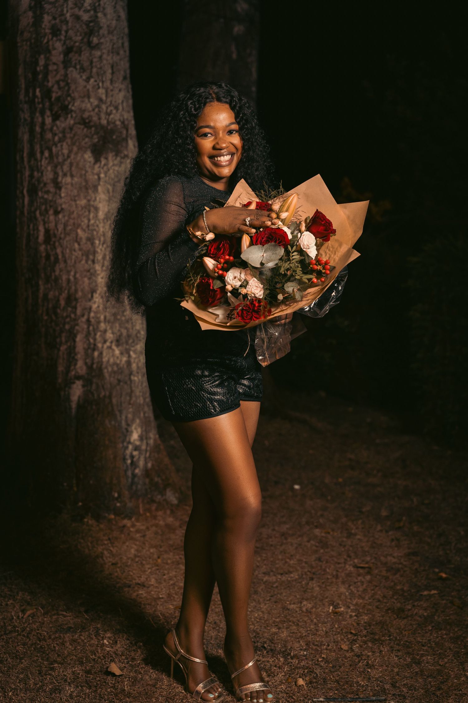 A person in a dark dress holds a beautiful floral bouquet while standing in a dimly lit forest setting.