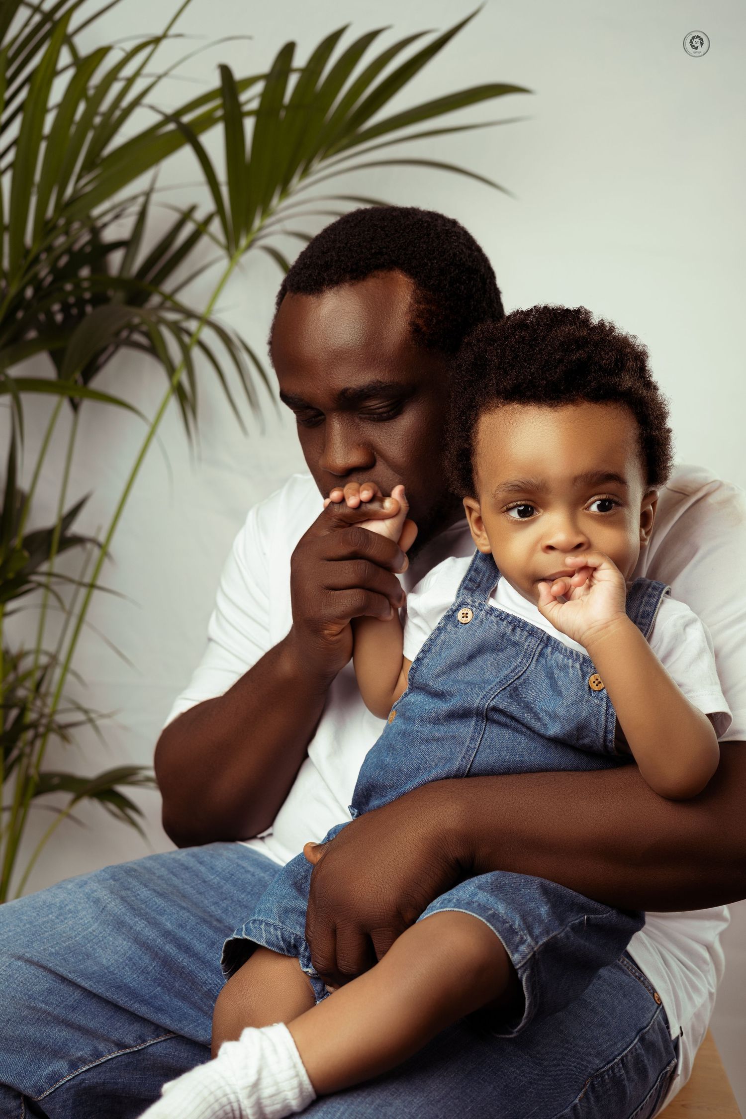 A parent and child share a tender moment together while sitting on a white couch with tropical plants in the background.