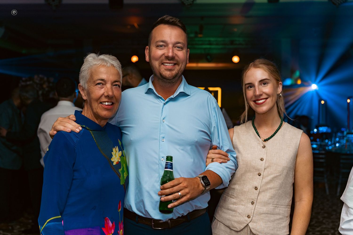 Three people smile together at an evening event while one holds a beer bottle in a blue-lit venue.