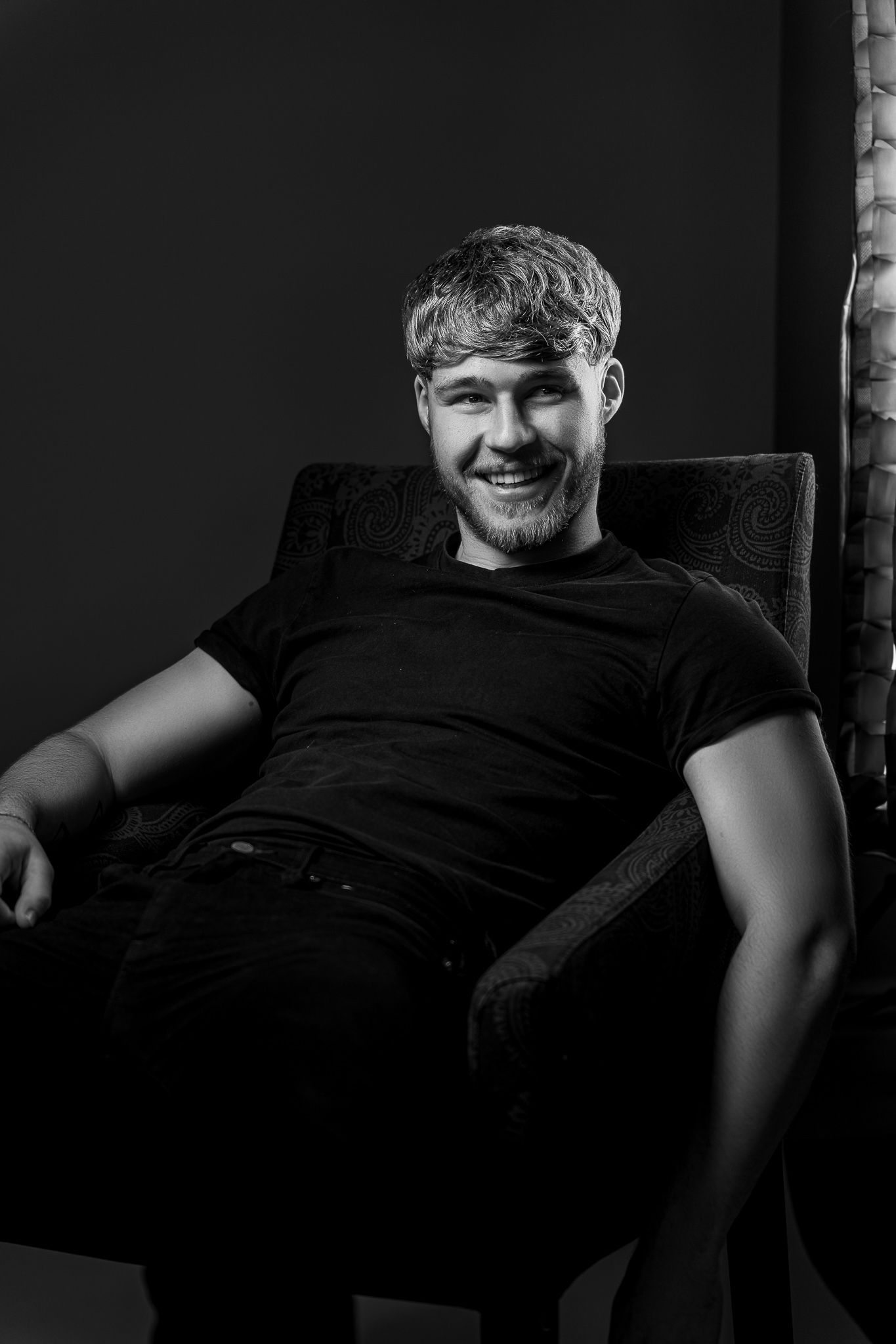Black and white portrait of person sitting in armchair wearing dark t-shirt and smiling at camera.