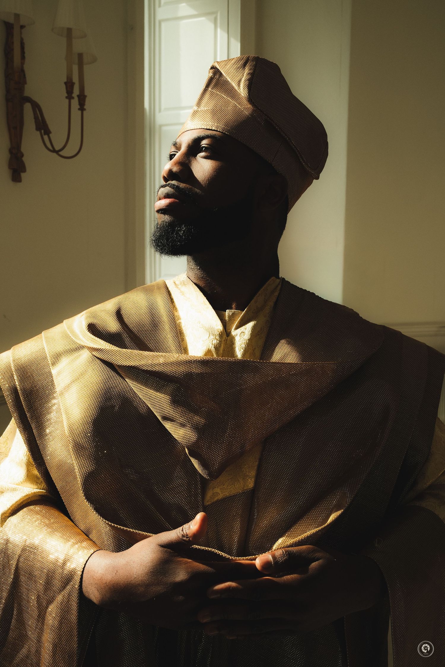Man in golden African traditional attire and cap, hands clasped, gazing upward in warm sunlight indoors.