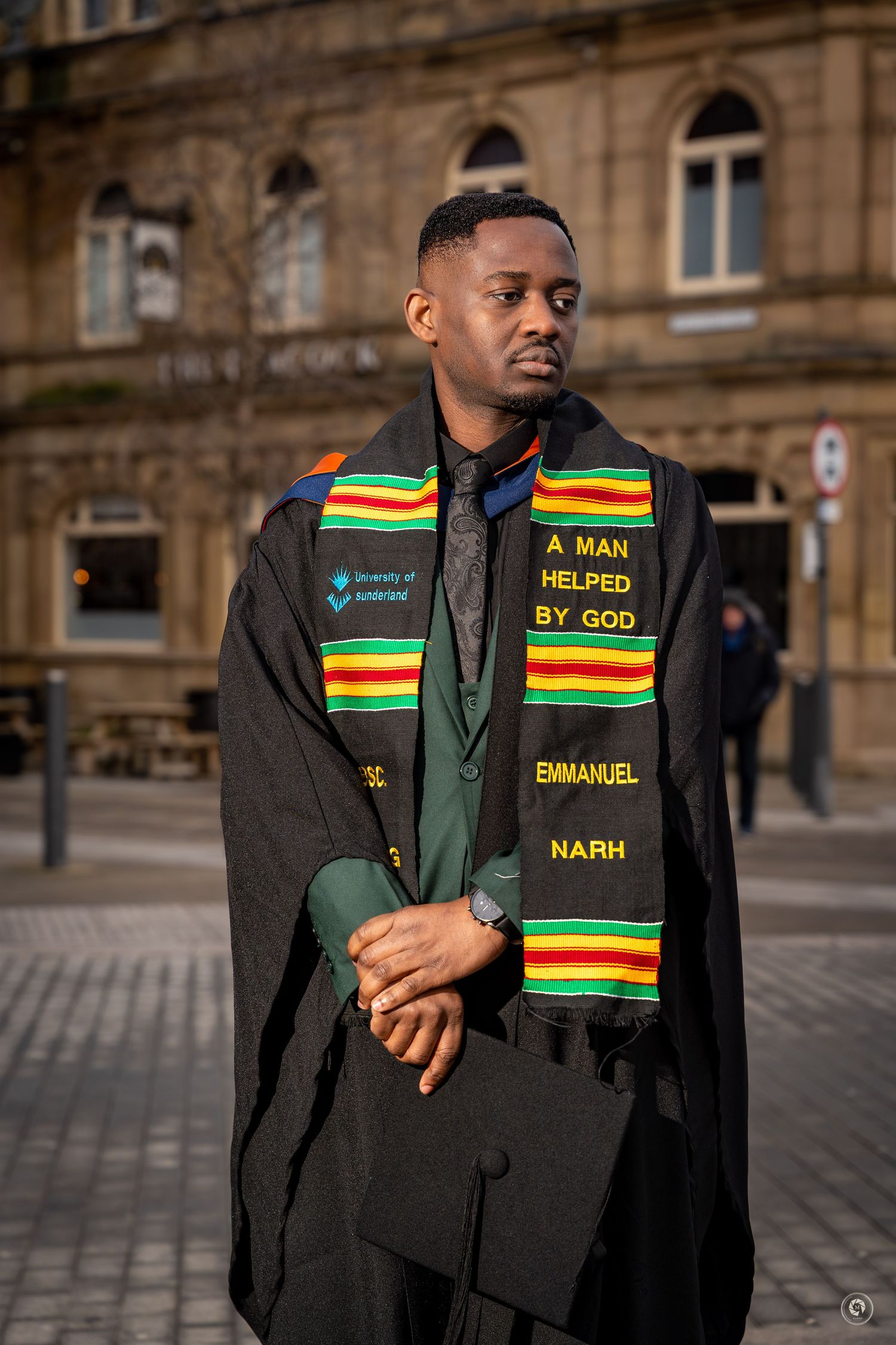 Graduate wearing black academic gown with colorful kente stole stands against stone building backdrop.