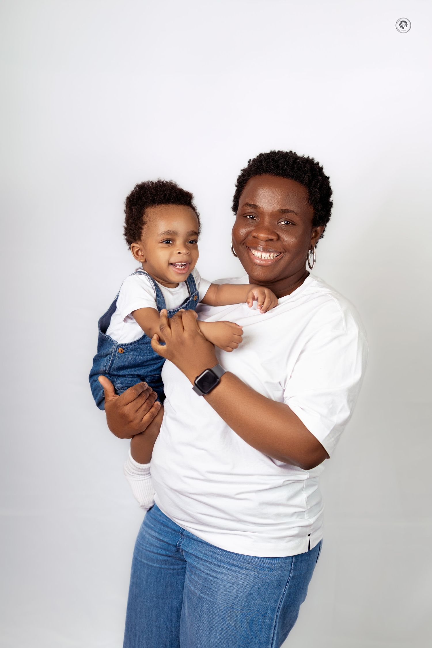 A parent in a white t-shirt and jeans holds and plays with a toddler wearing denim overalls against a white backdrop.