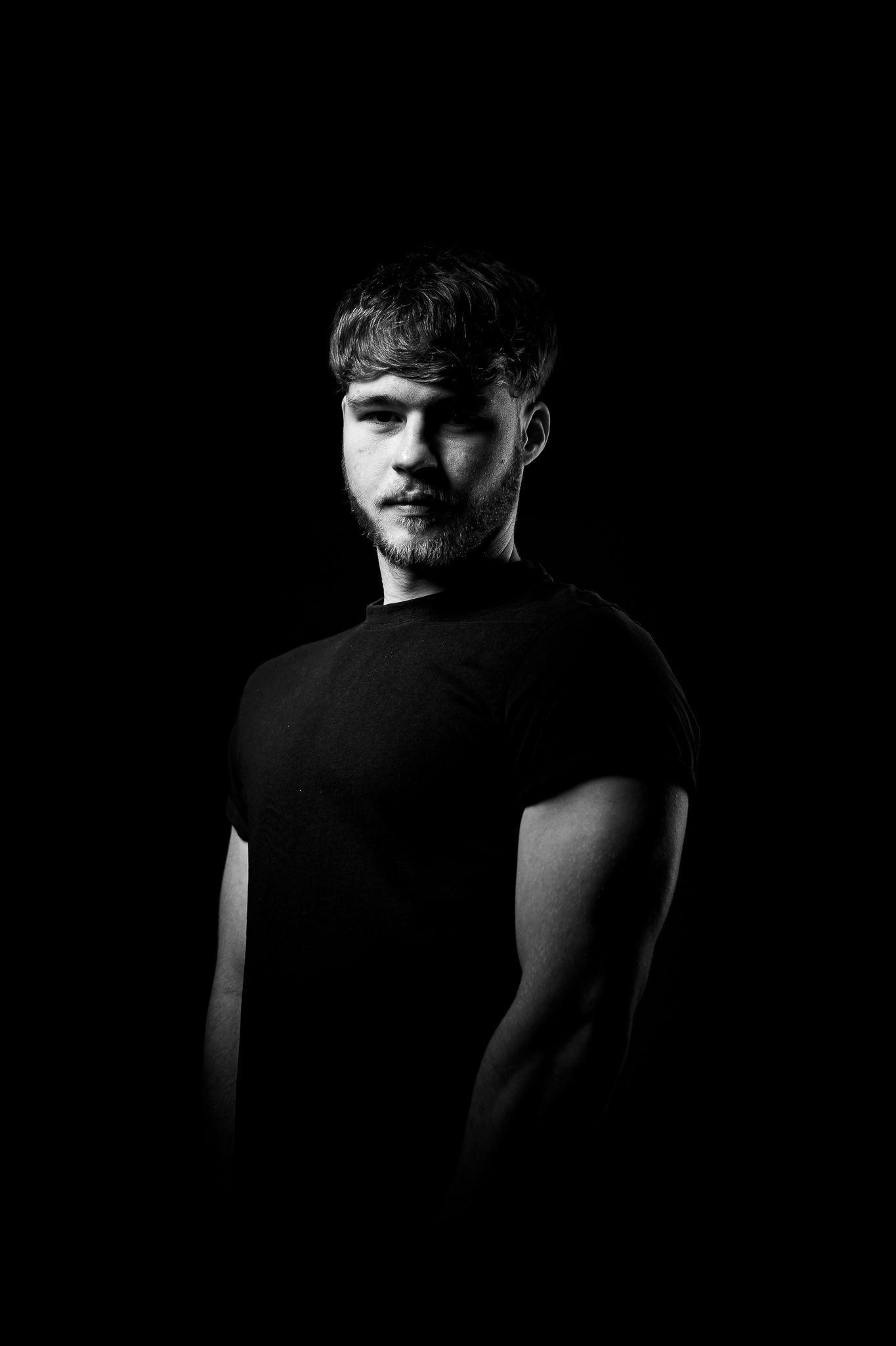 Dramatic black and white portrait in dark lighting features figure in t-shirt with dramatic shadows against black background.