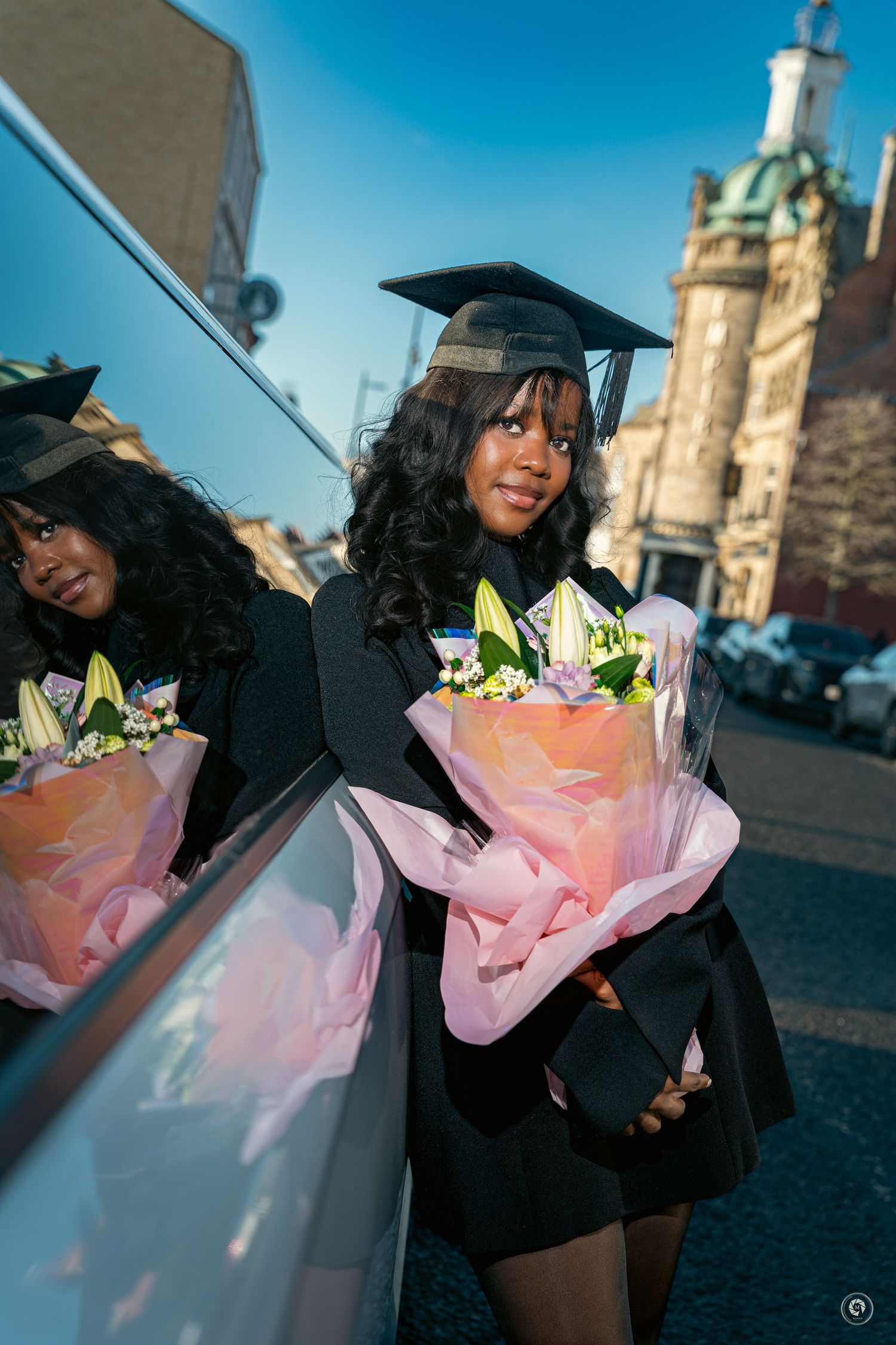 Graduate in black cap and gown holds pink-wrapped flower bouquet while reflecting in car window on sunny day.