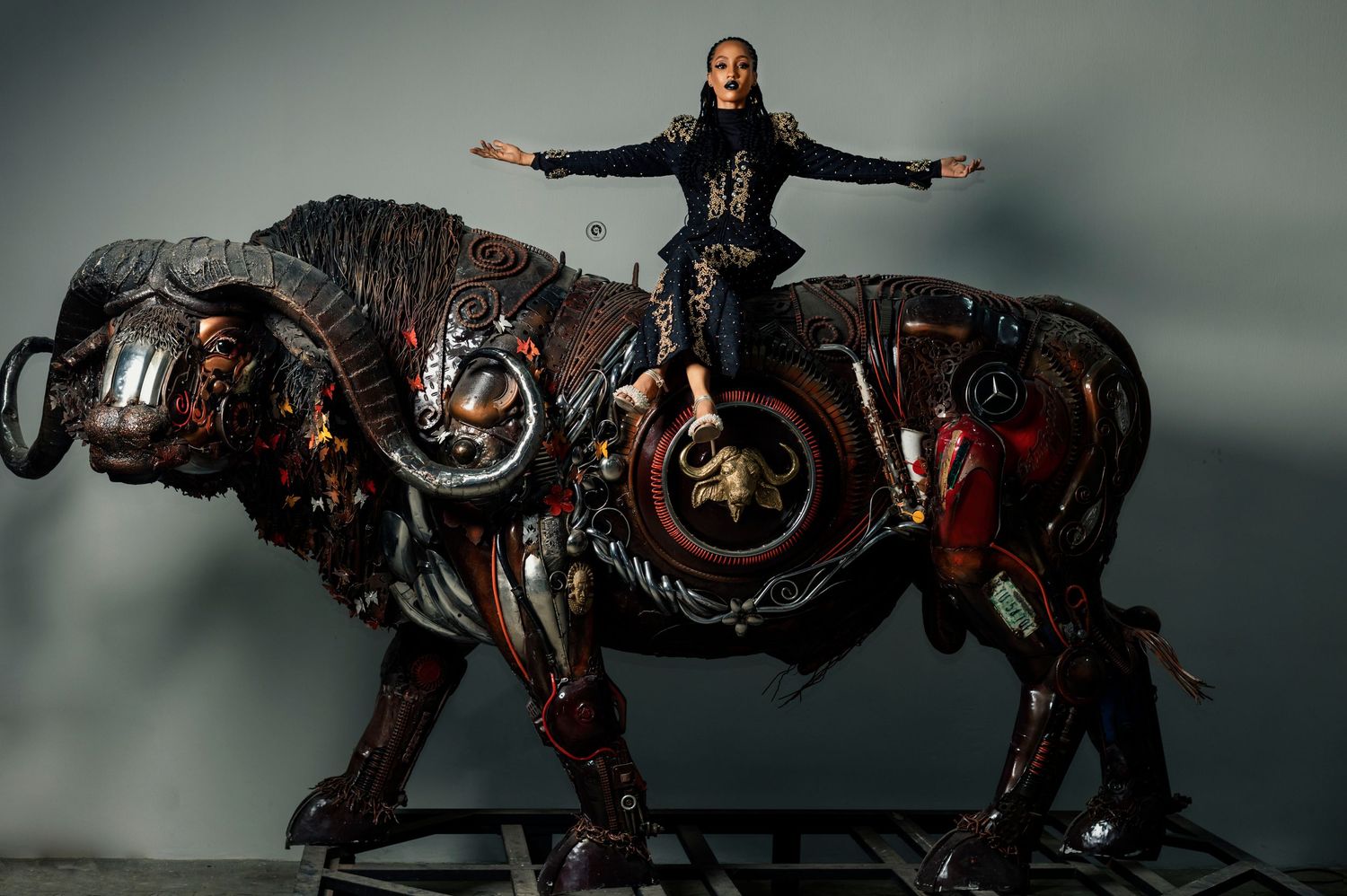 A dramatic steampunk sculpture of a mechanical bull with someone balancing on top in a dark studio setting.