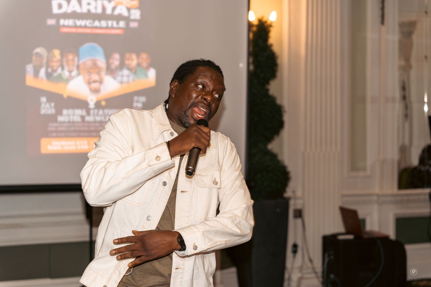 A performer in a white shirt holds a microphone while speaking at an event with a projection screen visible in background.