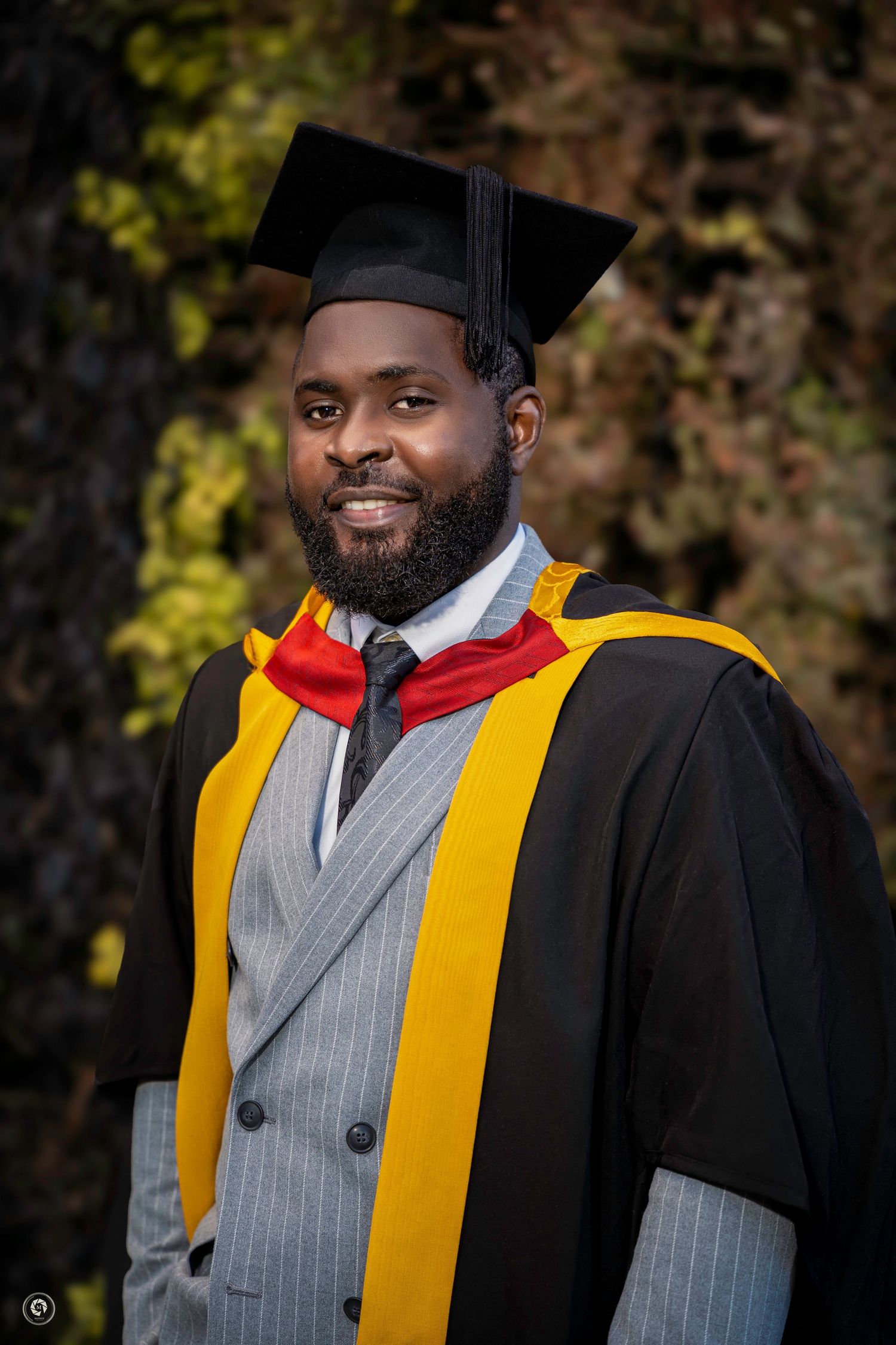 Graduate in academic regalia wearing black gown with gold stole and gray vest smiles during outdoor graduation photoshoot.