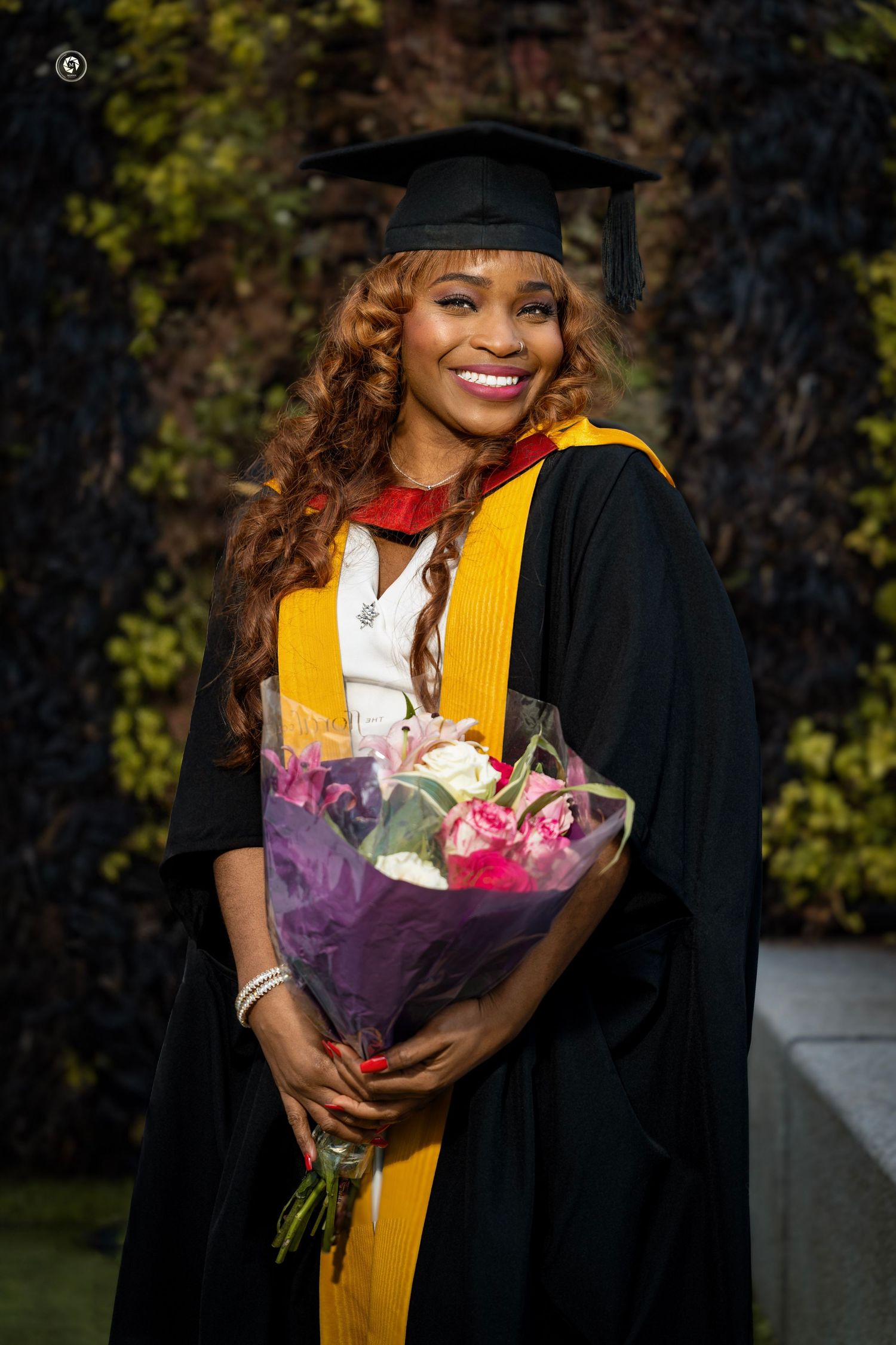 Graduate wearing yellow stole and black gown holds purple flower bouquet during outdoor graduation photo session.