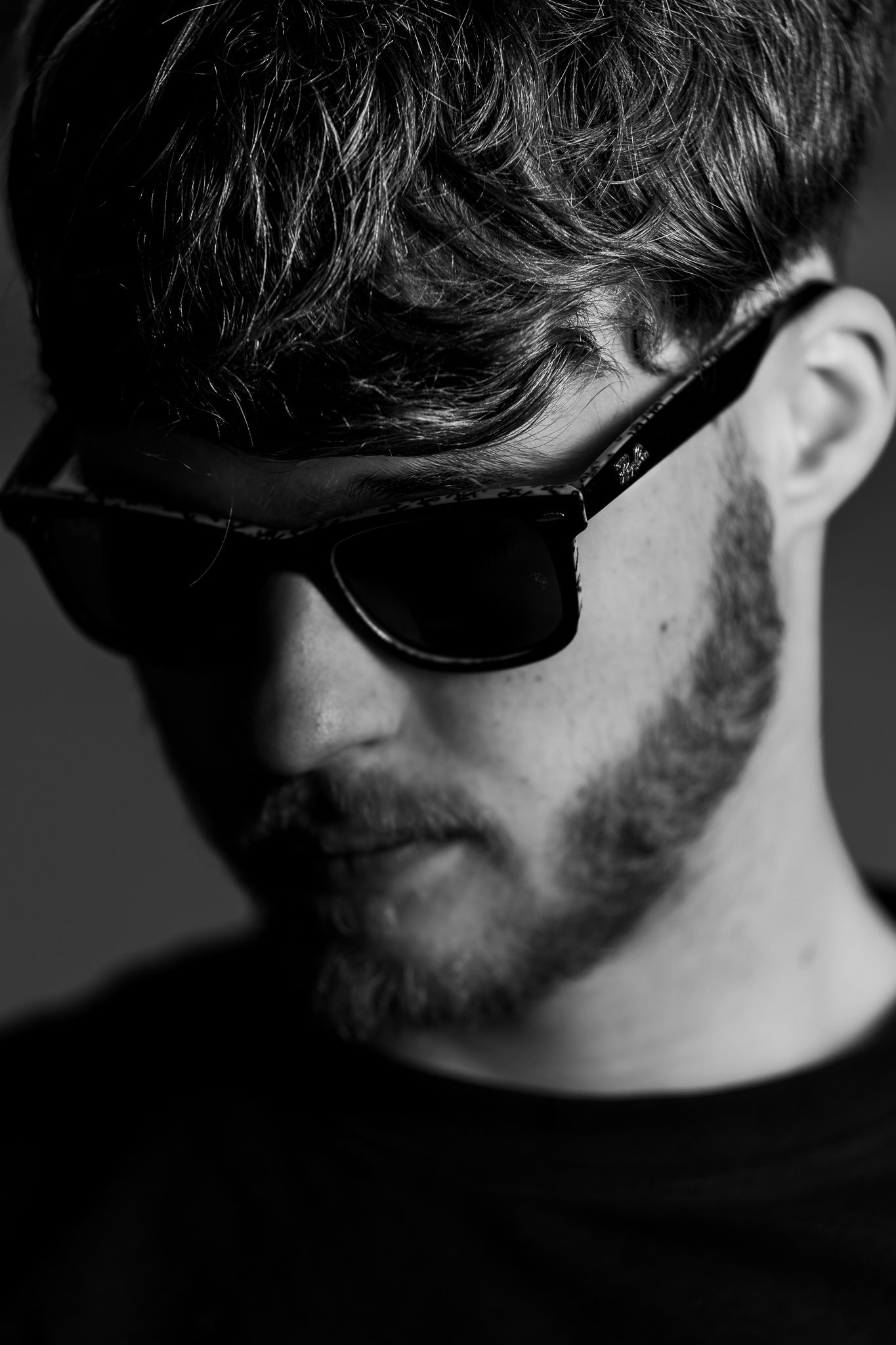 Black and white close-up portrait of person wearing sunglasses with dark tousled hair and facial hair against dark background.