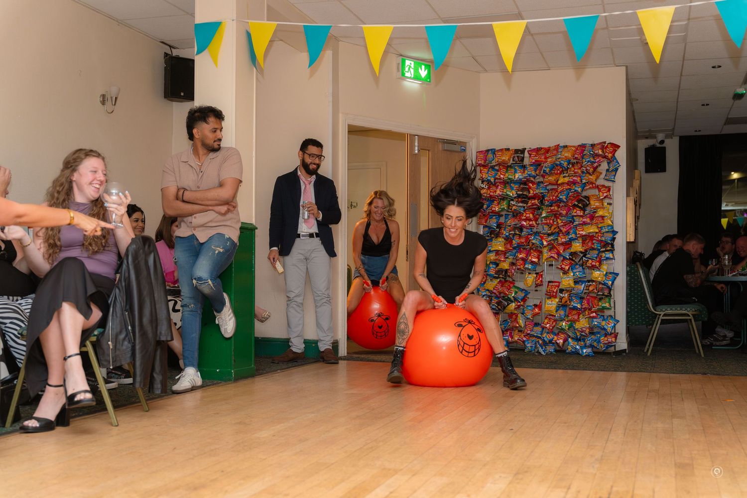People gather in a room decorated with colorful bunting flags while two individuals bounce on orange exercise balls.