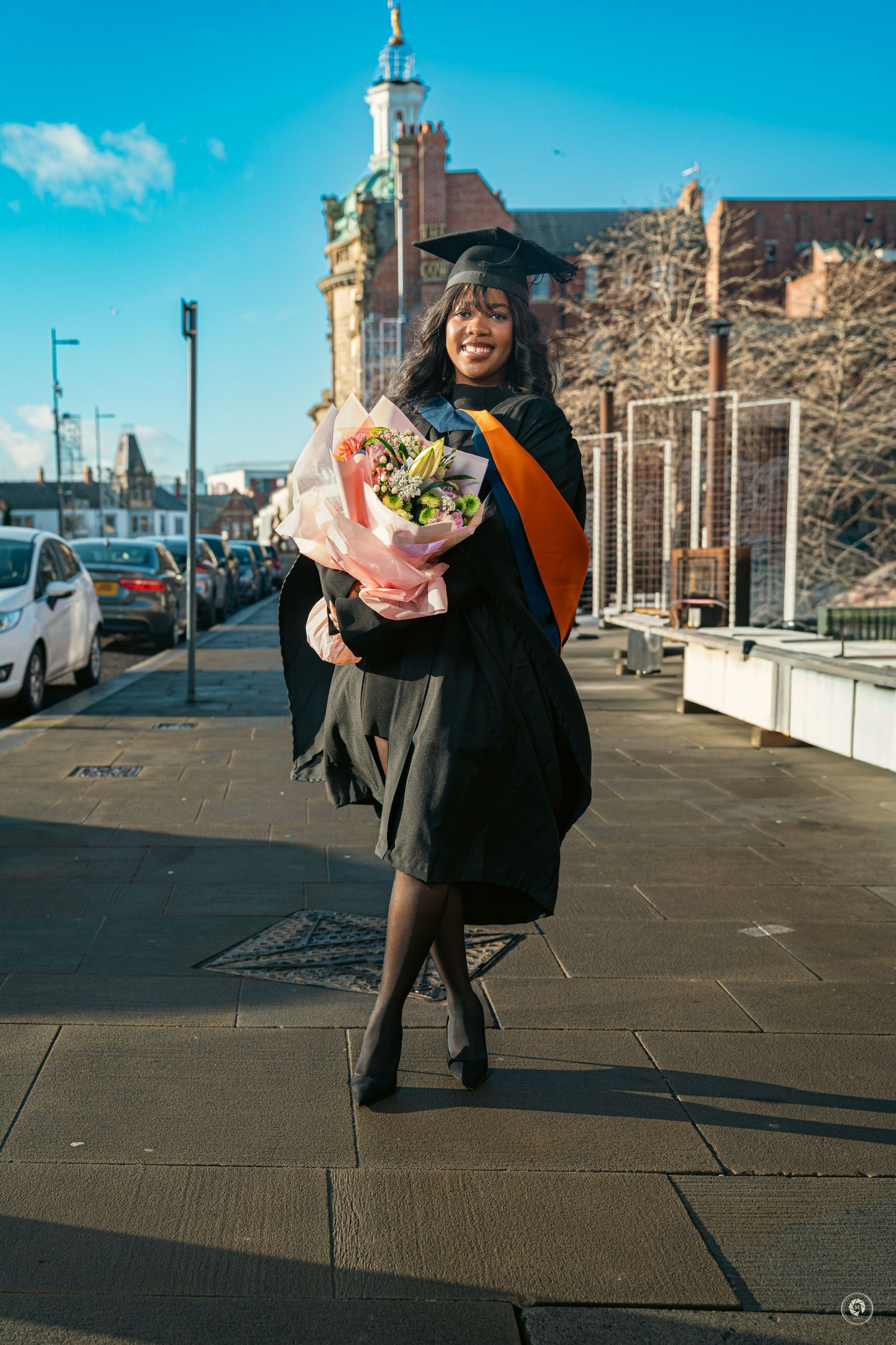 Someone in a black coat holds a bouquet of flowers while walking along a sunny seaside promenade.