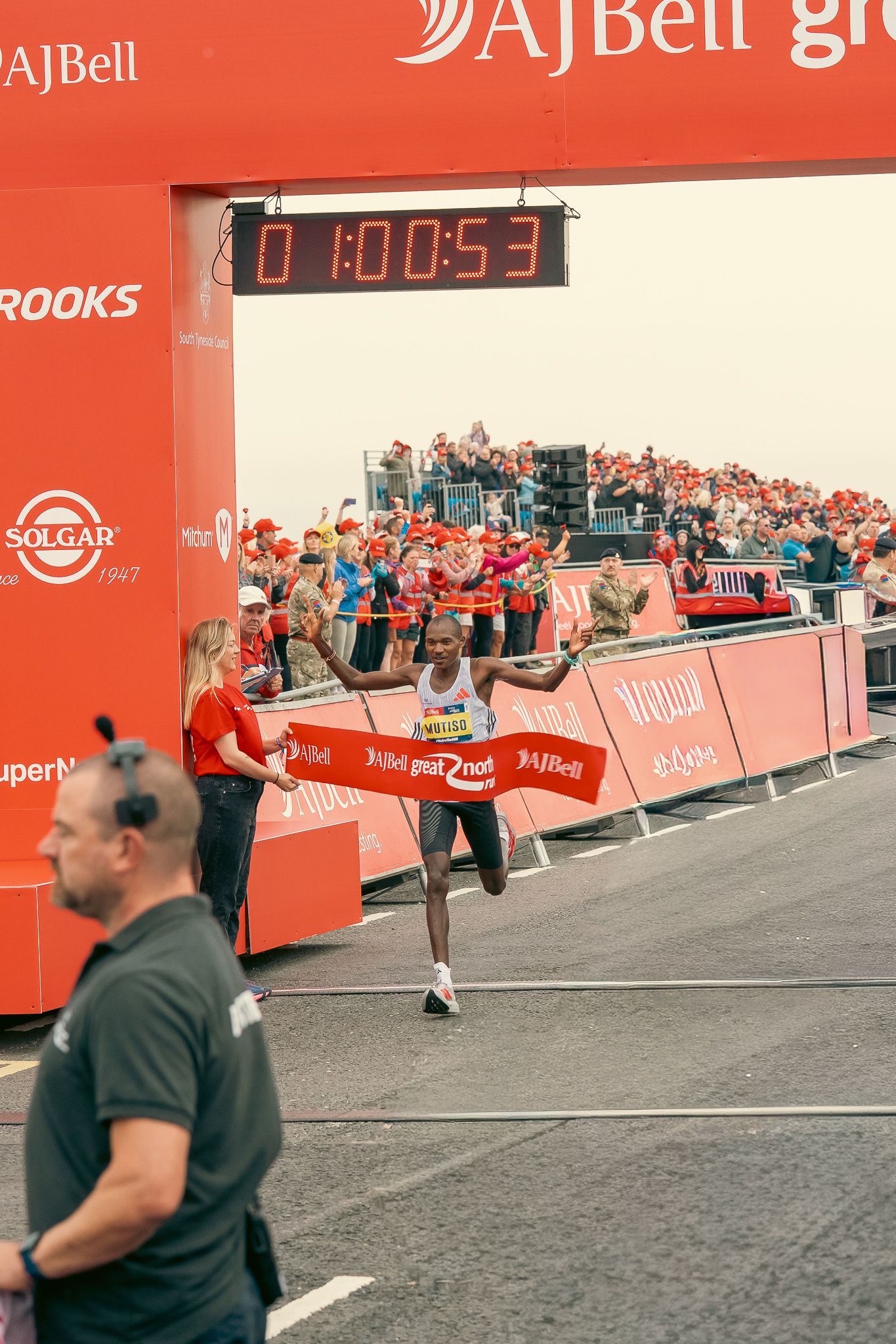 Runners pass by red AJBell sponsor banners and crowd barriers during a race event with spectators in background.