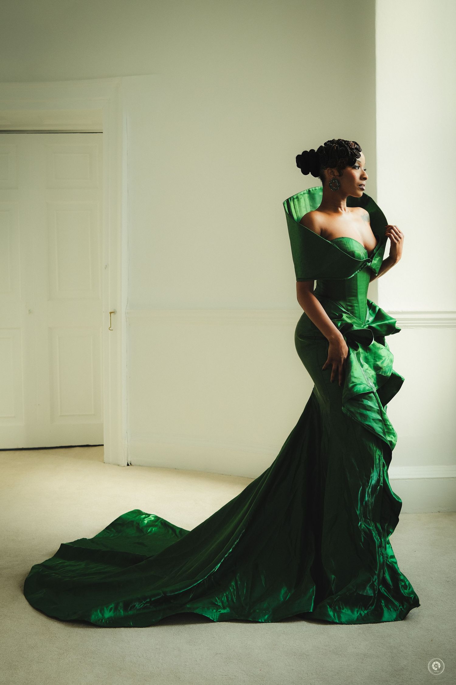 A woman in an elegant emerald green satin gown with dramatic ruffles and a long train poses gracefully indoors.