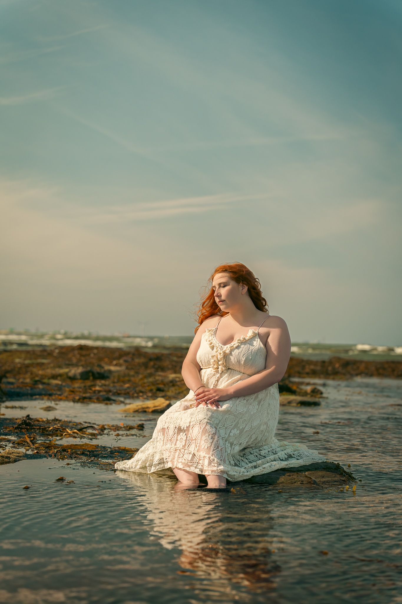 Woman in white lace dress sits gracefully on coastal rocks as gentle waves wash around her during golden hour.