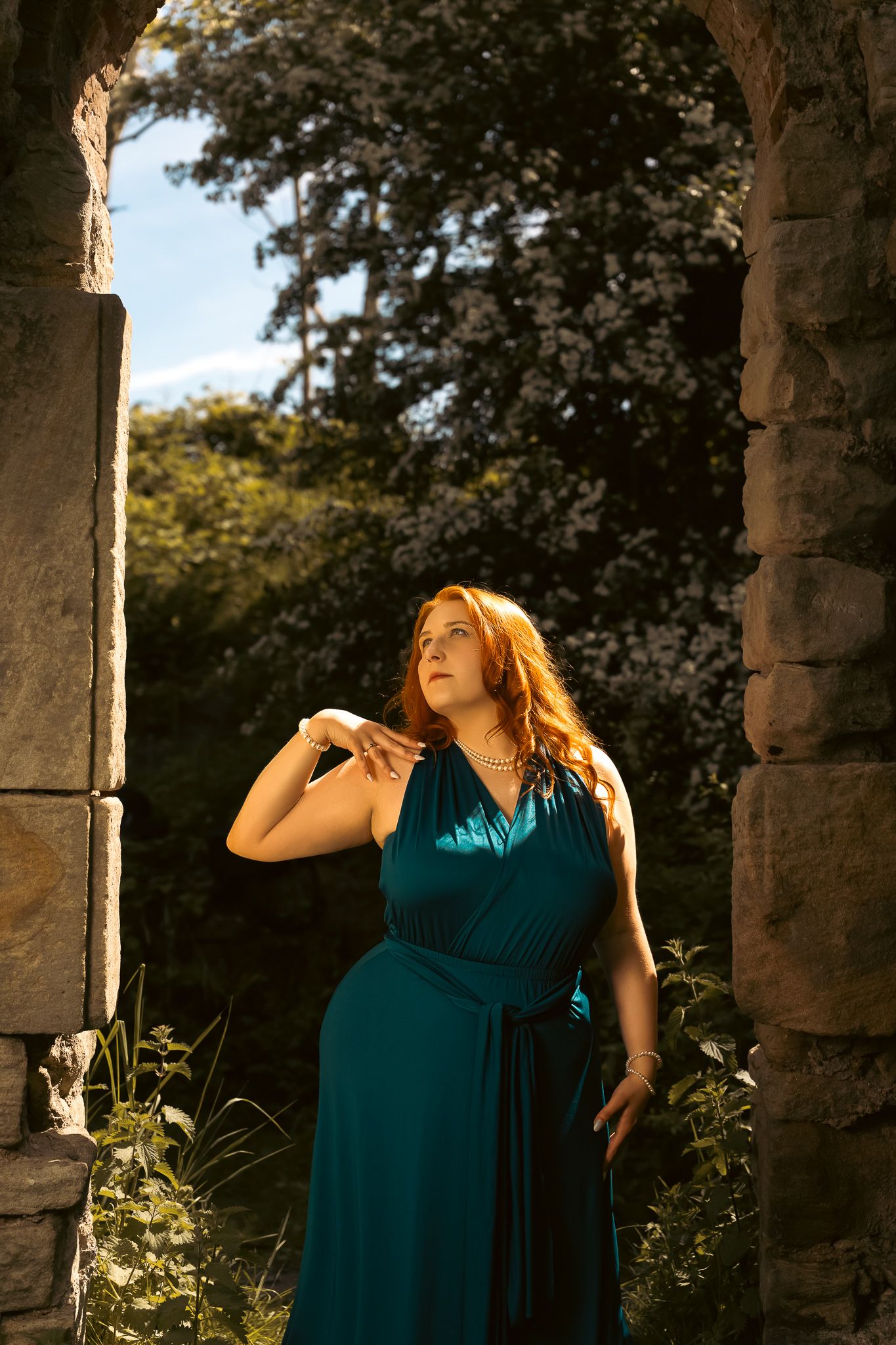 A beautiful teal evening dress glows in golden sunlight through stone archway surrounded by lush greenery.