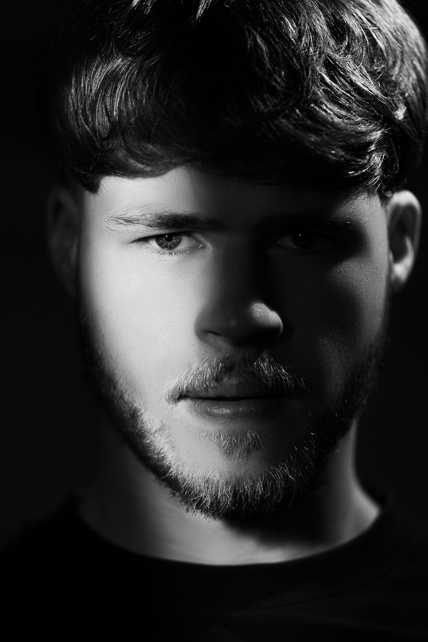 Black and white dramatic portrait with strong side lighting emphasizing facial features and beard in shadow.