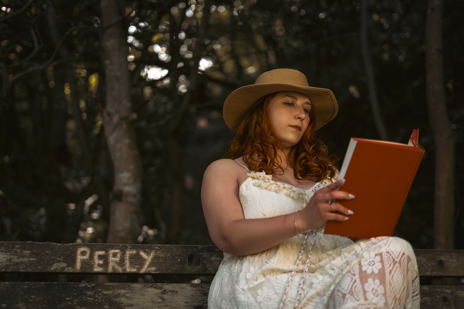 Person in white dress and brown hat reads book while sitting on wooden bench in moody forest setting.