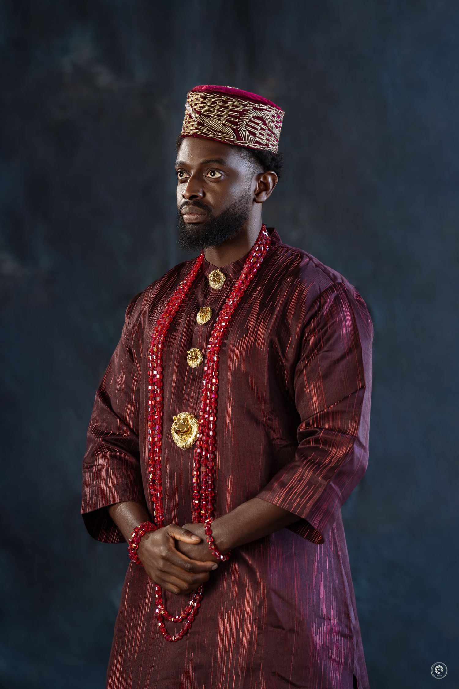 Man in traditional African attire with red beaded necklace, gold medallion, and embroidered cap poses formally.