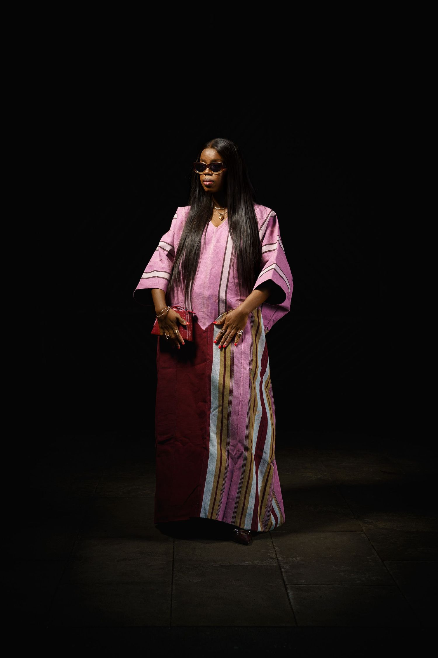 Woman in pink striped African dress and sunglasses poses dramatically against dark background.