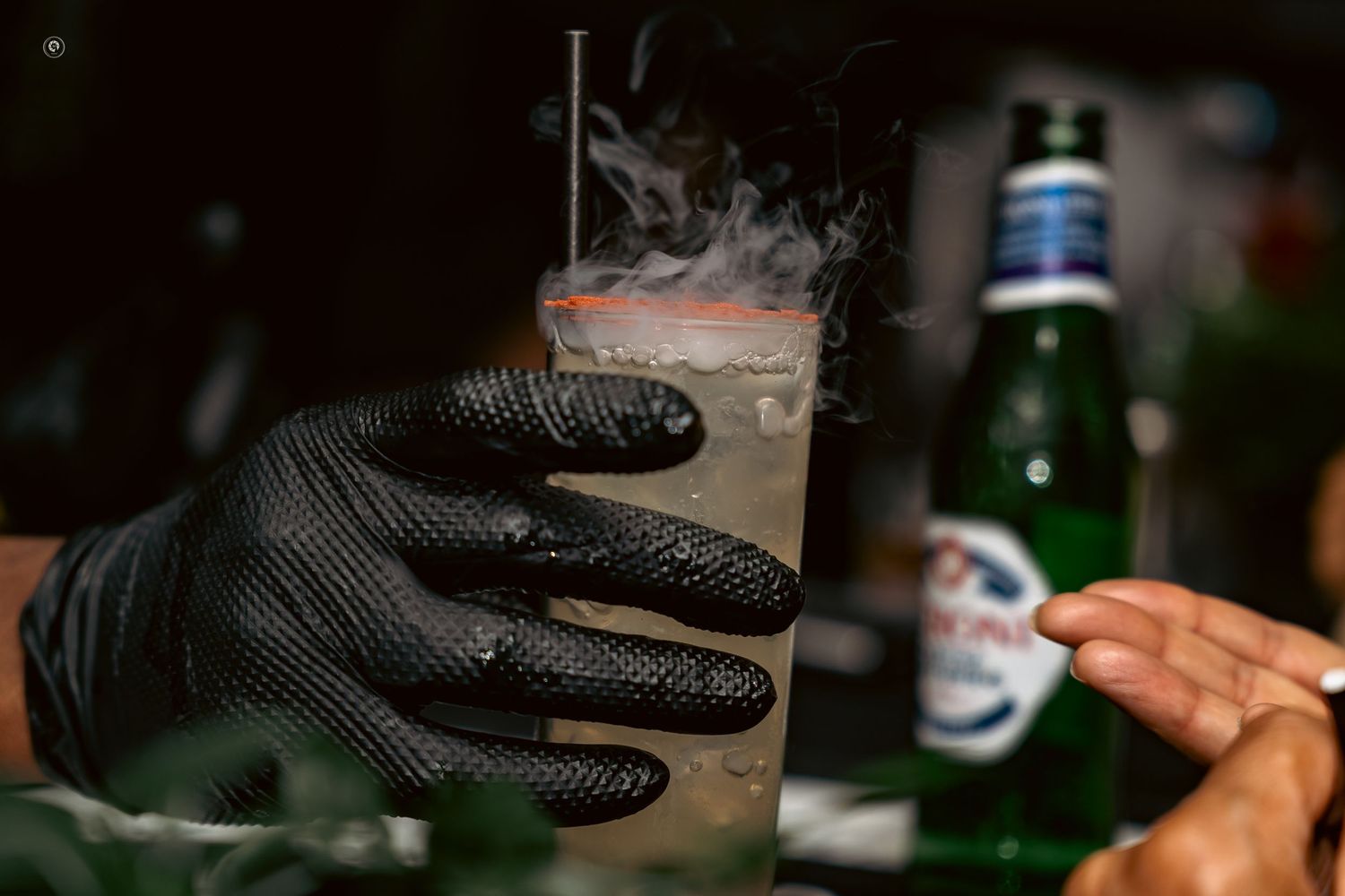 A hand wearing black gloves pours a clear liquid from a bottle into a glass container.
