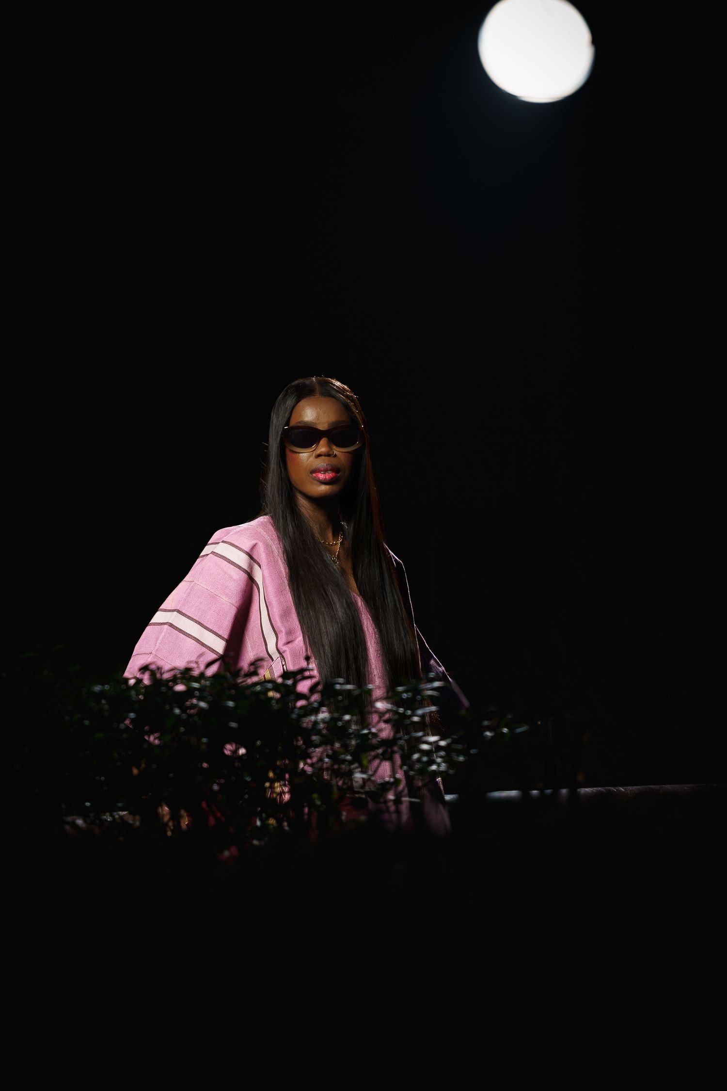 A Black woman in a pink jacket and sunglasses stands in dramatic dark lighting with a glowing orb above.