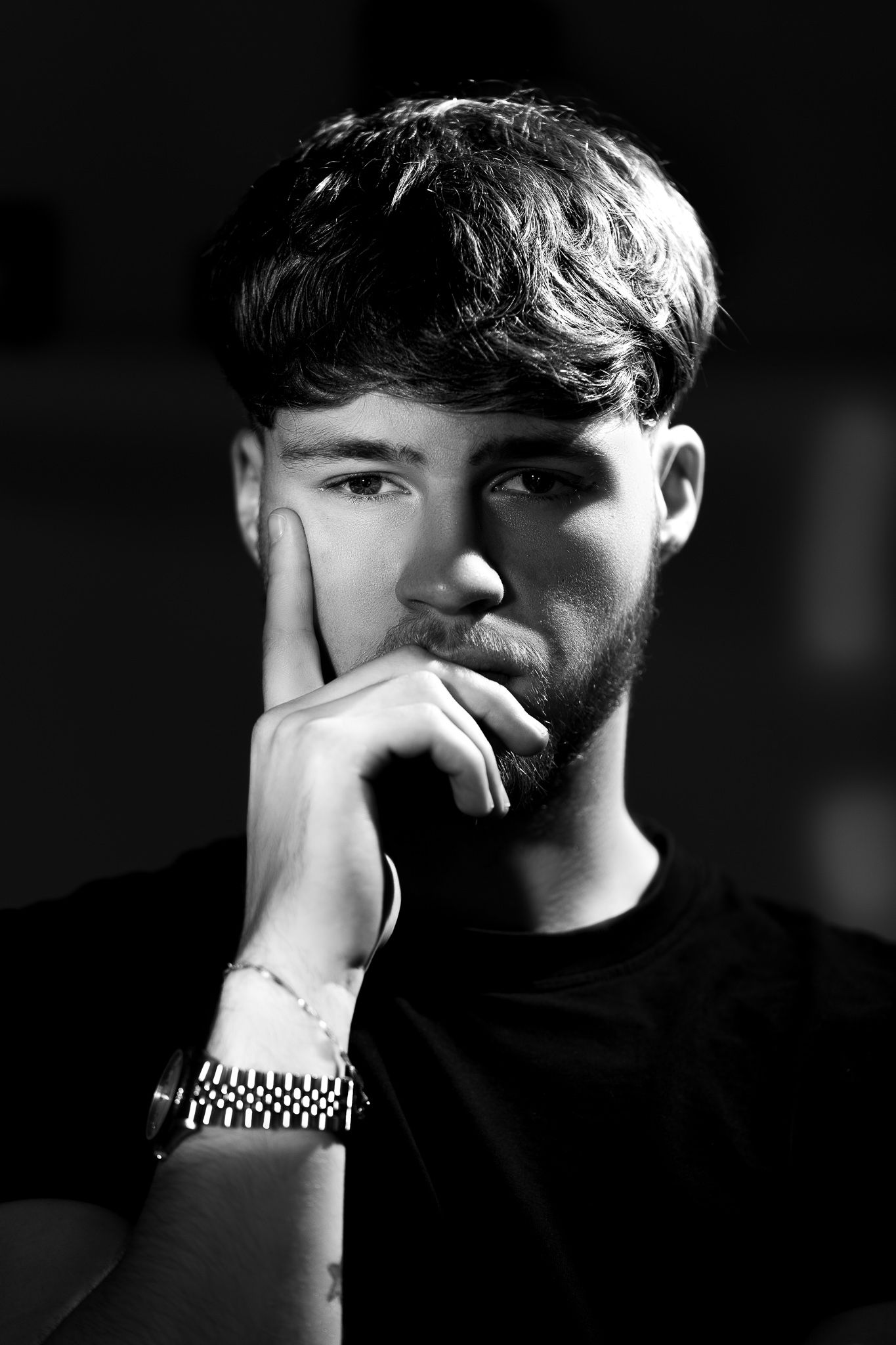 A black and white portrait of a person with short hair holding their hand to their face while wearing a watch and dark clothing.