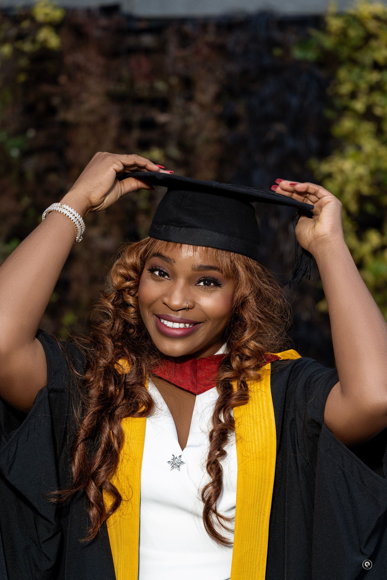 Graduate wearing academic regalia with black cap, white dress, yellow stole and red hood smiles during graduation photo.