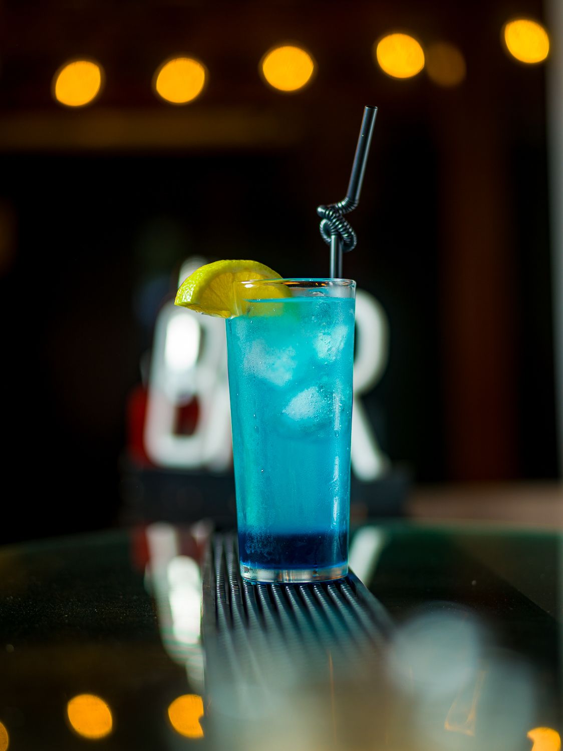 A vibrant blue electric colored cocktail sits in a clear glass with tropical garnish and bokeh lights in the background.