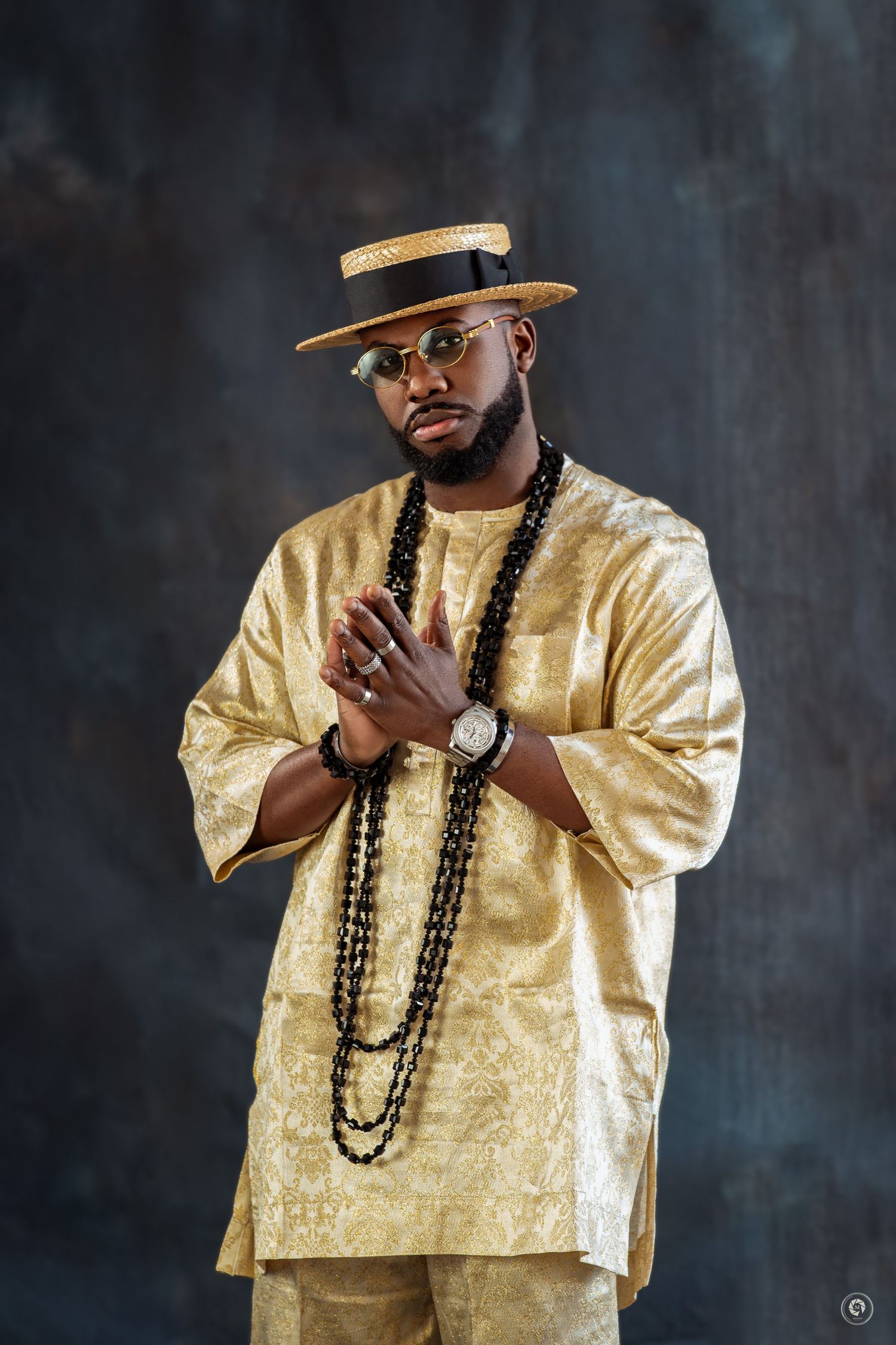 Man in gold African attire, beaded necklace, straw hat, and glasses poses with hands clasped in studio portrait.